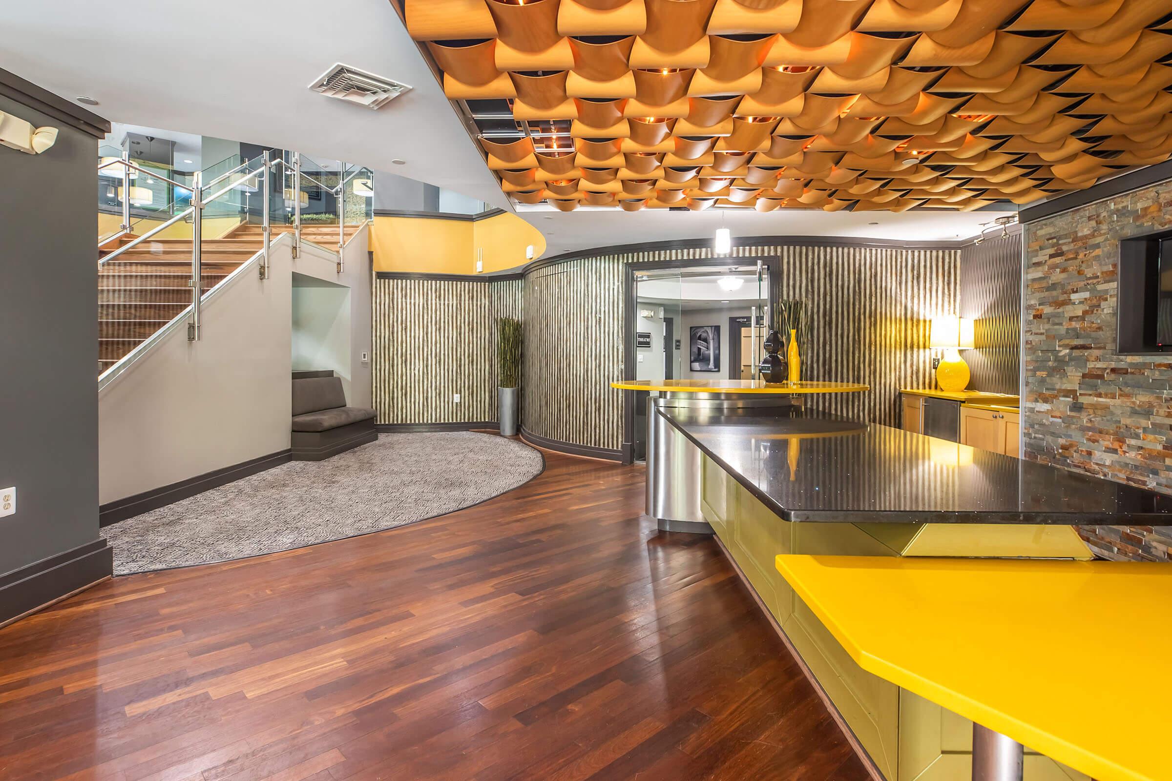 Interior view of a modern lobby featuring a curved wooden accent wall, a sleek countertop with a yellow finish, and wooden flooring. The ceiling has an artistic design, and a staircase leads to a second level. The space includes a cozy seating area and contemporary decor elements.