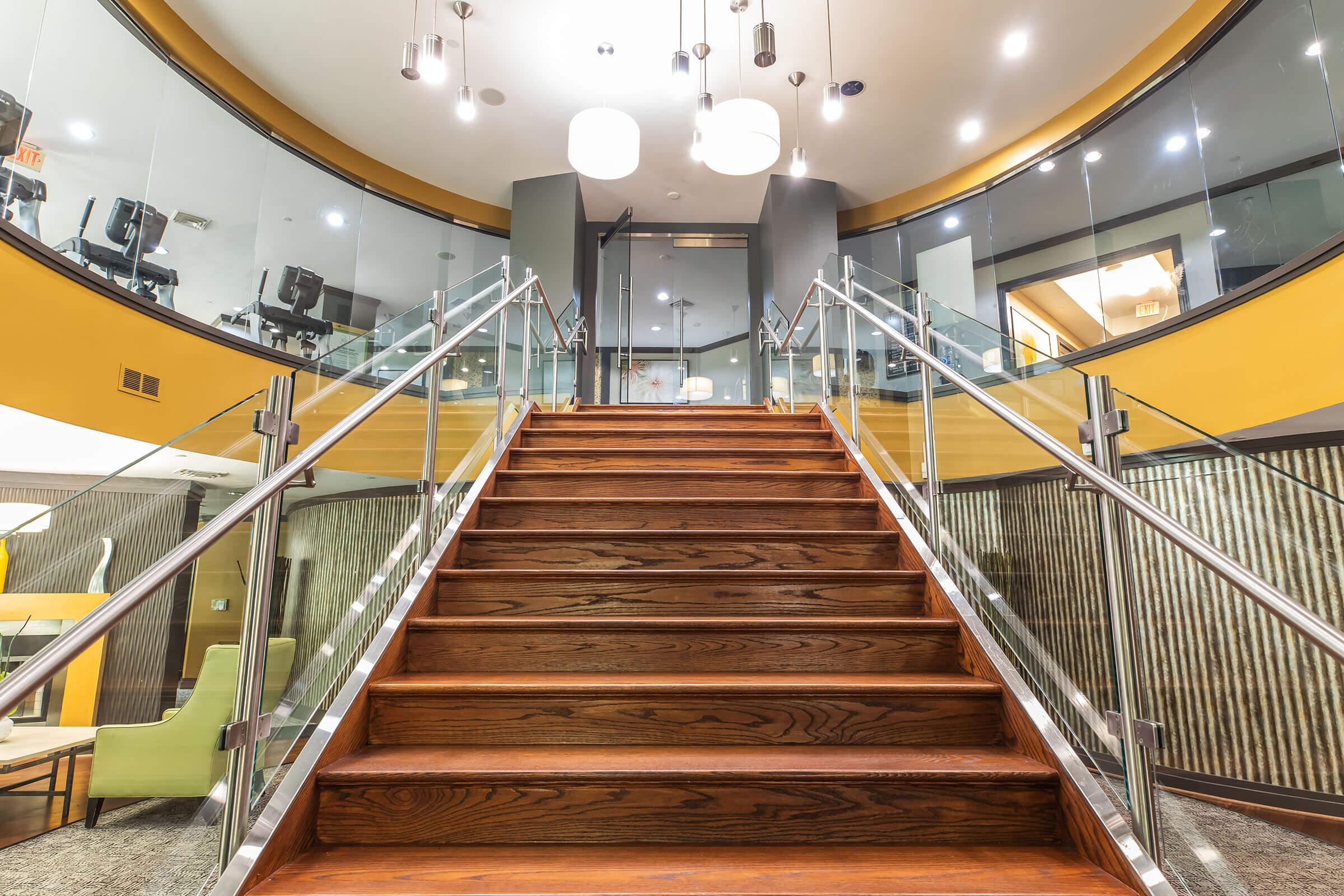 A modern indoor staircase with wooden steps and a glass railing leads to a second level. The surrounding area features contemporary lighting fixtures and a stylish, open design, showcasing a welcoming atmosphere in a well-lit space.