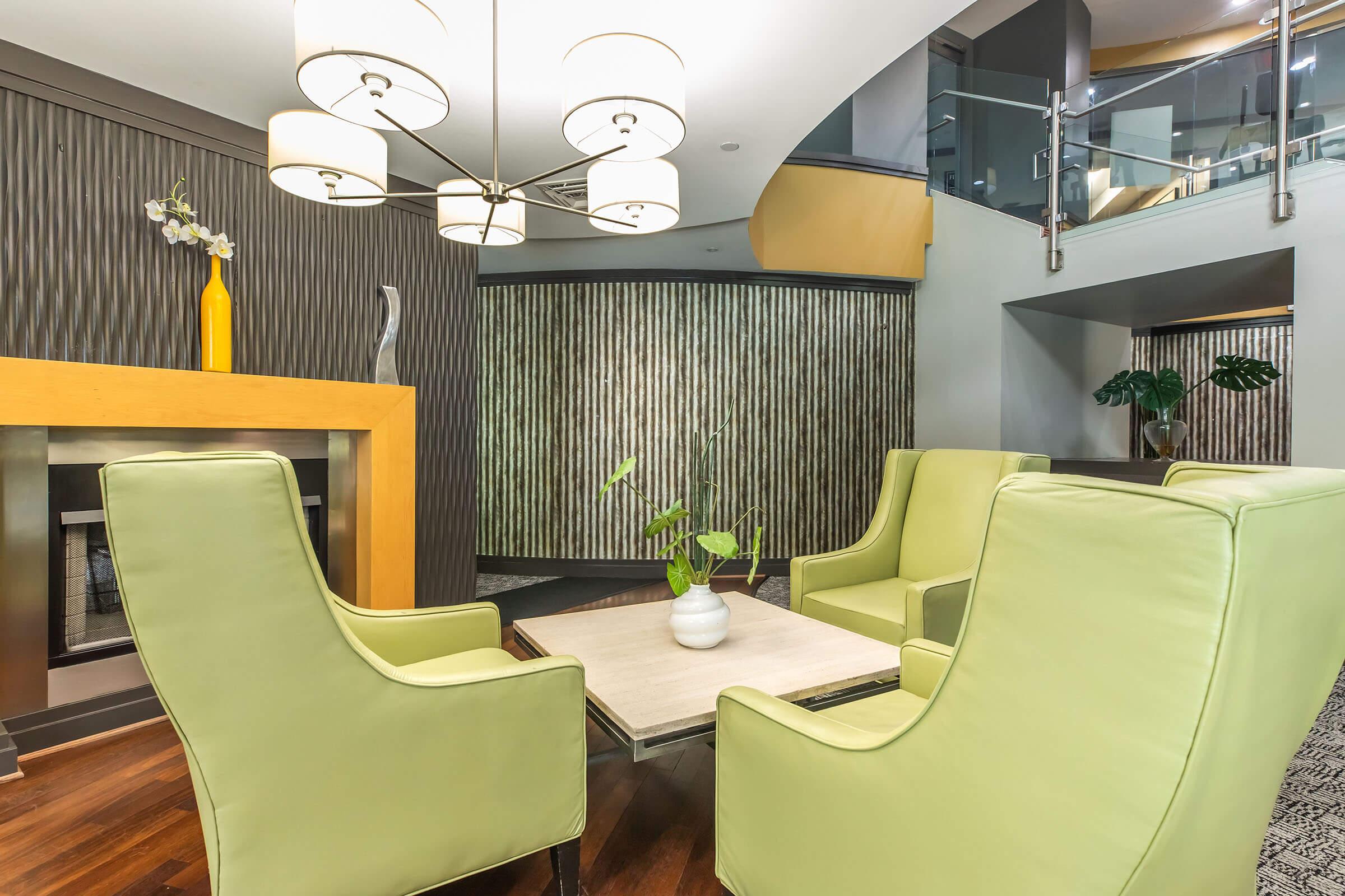 A modern lounge area featuring two light green armchairs and a small coffee table. A yellow vase with flowers sits on the table. The backdrop includes a textured wall and a decorative feature behind the seating. Soft lighting is provided by a stylish chandelier above.