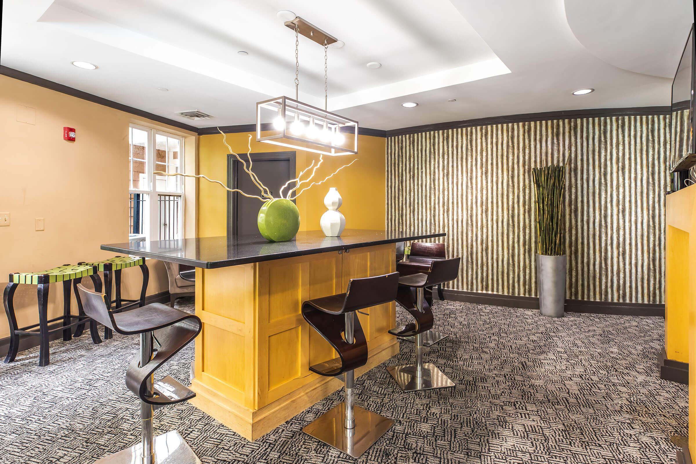 A modern kitchen or bar area featuring a sleek countertop with a decorative green vase and a unique white sculpture. Stylish bar stools line the counter. The walls are painted a warm yellow with a textured accent wall, and there is a touch of greenery in a tall vase. The flooring has a patterned carpet design.