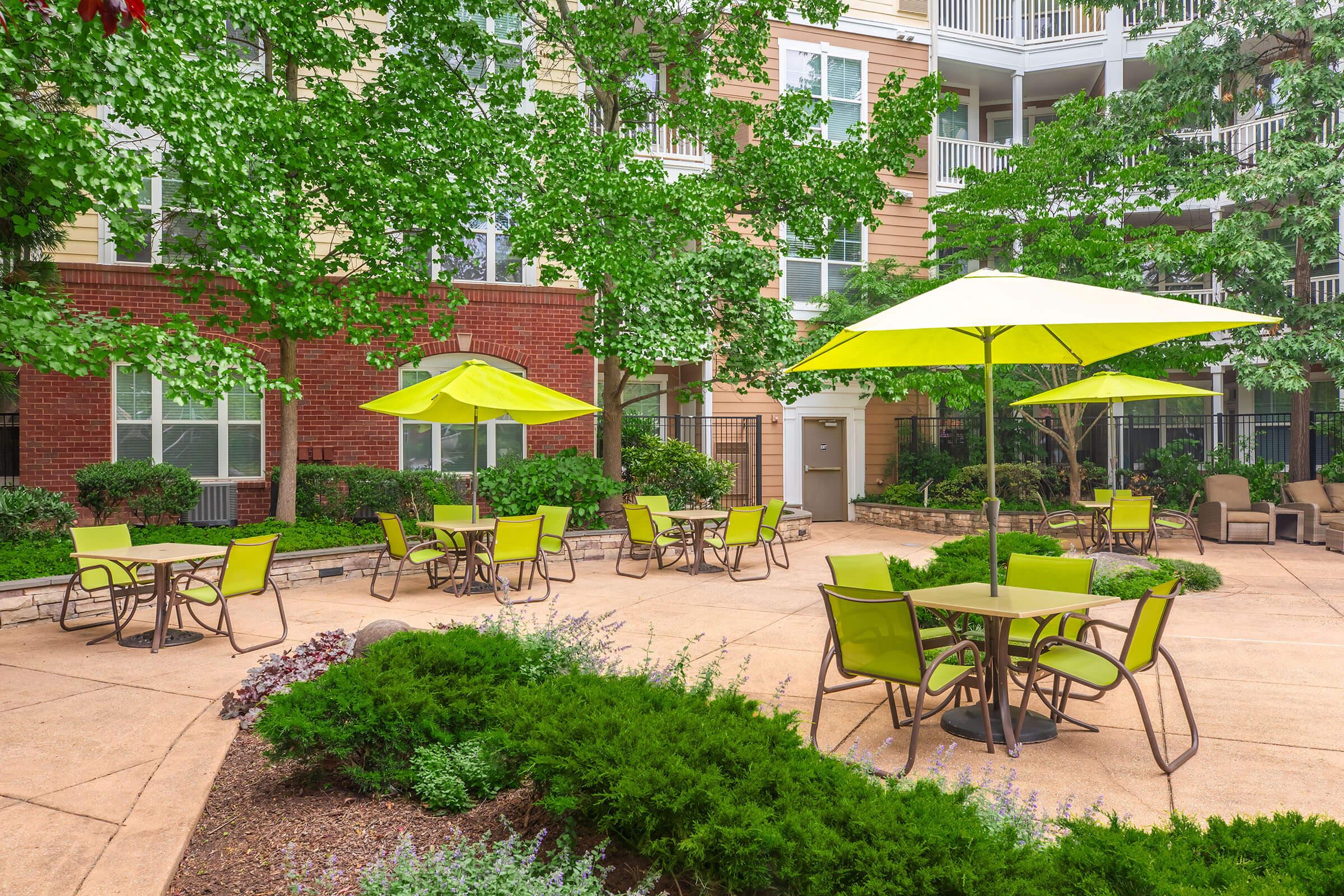 A sunny outdoor area with green chairs and tables, shaded by bright yellow umbrellas. Surrounding greenery includes bushes and trees, creating a pleasant and inviting atmosphere. The backdrop features a multi-story building with balconies. Ideal for relaxation and social gatherings.