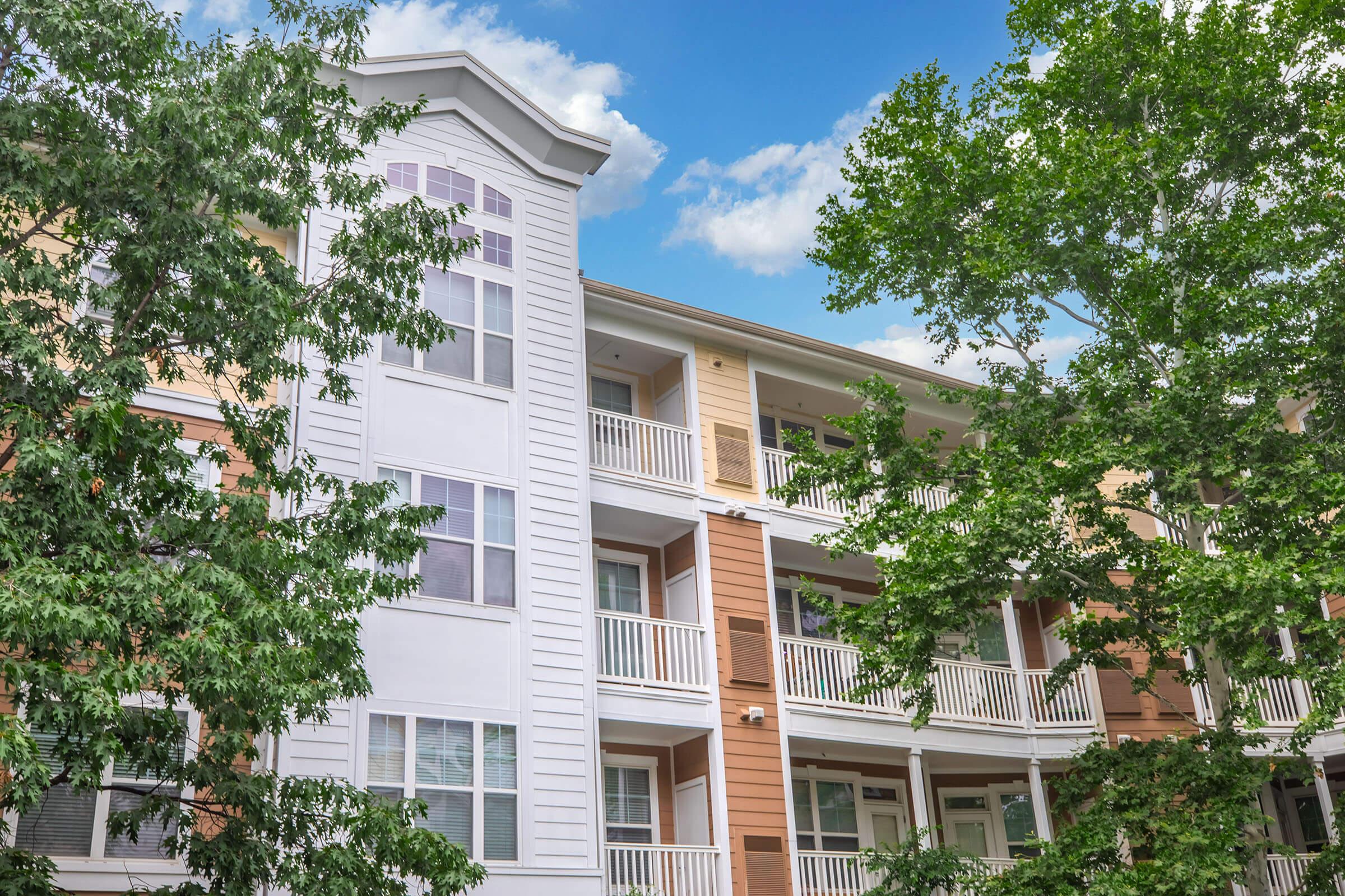 A modern multi-story apartment building with balconies, featuring a combination of white, light brown, and dark brown siding. Lush green trees partially frame the building against a bright blue sky with scattered clouds. The architecture reflects contemporary design with ample windows.