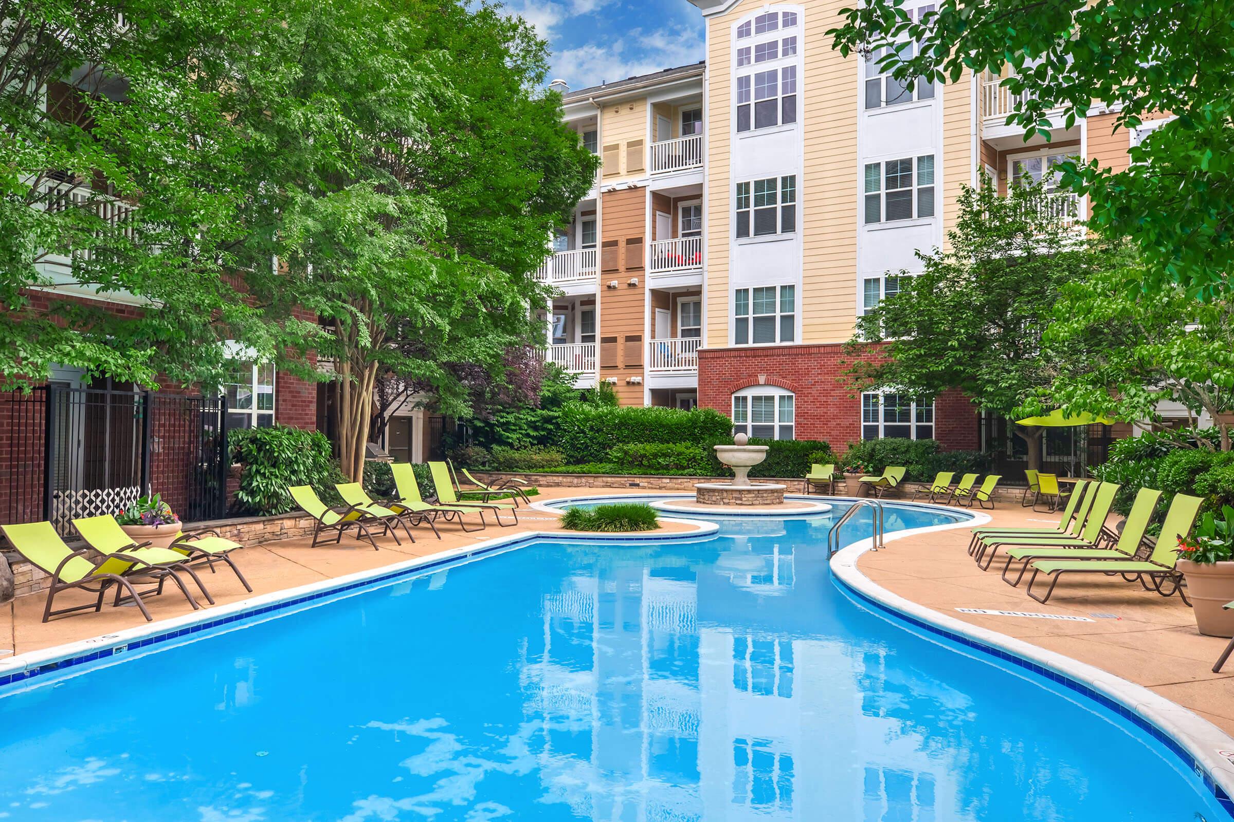 A serene swimming pool surrounded by lush greenery and comfortable lounge chairs. The pool features a gentle curve, with a small fountain at one end. Apartment buildings with large windows are visible in the background, creating a peaceful and inviting outdoor atmosphere.