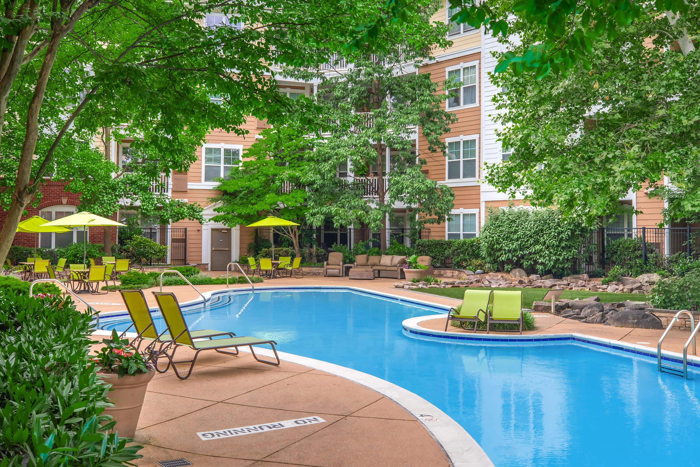 A tranquil swimming pool area surrounded by lush greenery and trees. Sun loungers with bright green cushions are positioned around the pool, with yellow umbrellas providing shade. The pool features a clear blue water surface and a winding shape, set against a backdrop of residential buildings.