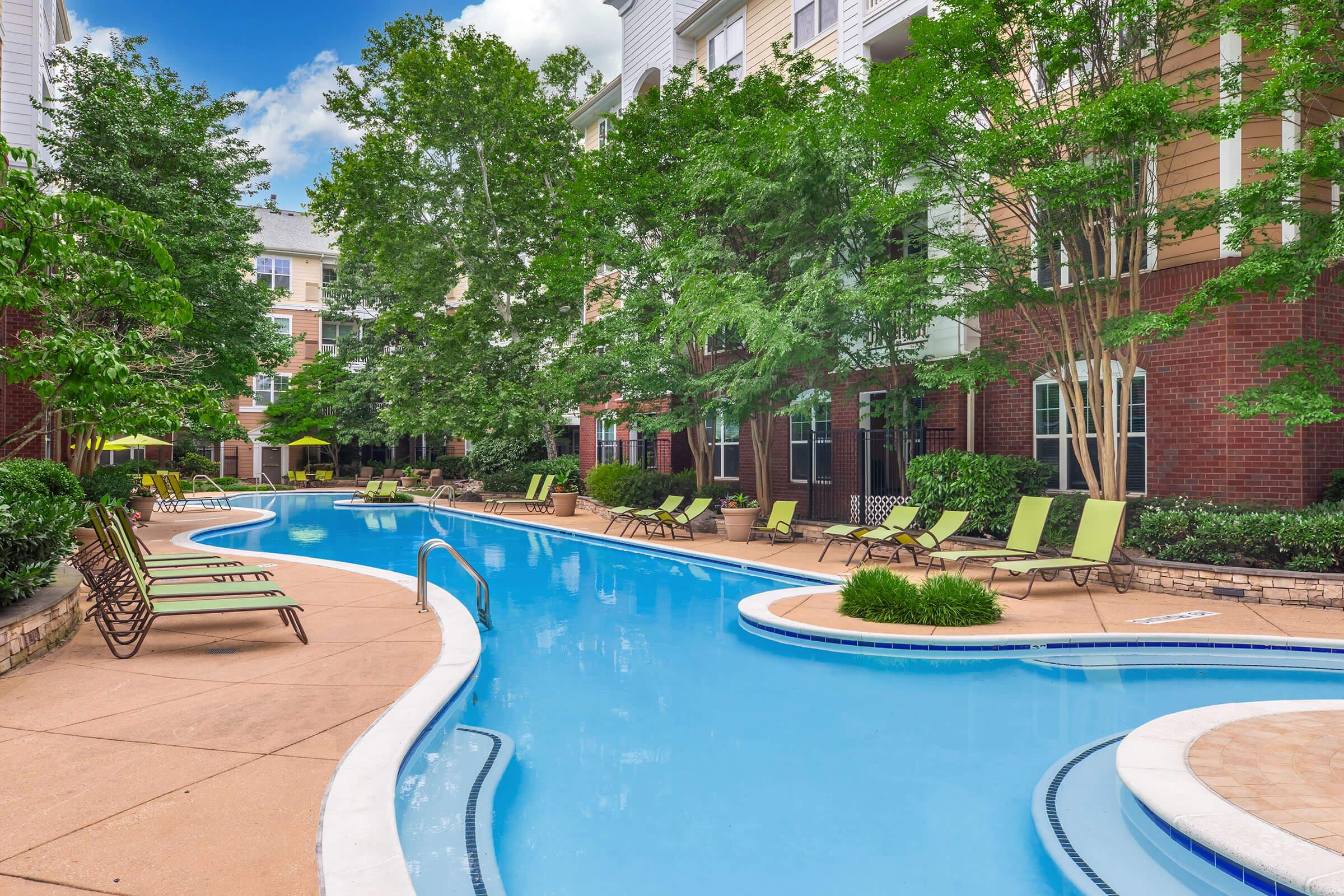 A serene outdoor pool area surrounded by lush greenery, featuring a winding pool with lounge chairs on the deck. The area is well-lit with sunny skies and a mix of residential buildings in the background, creating a relaxing atmosphere for leisure and recreation.