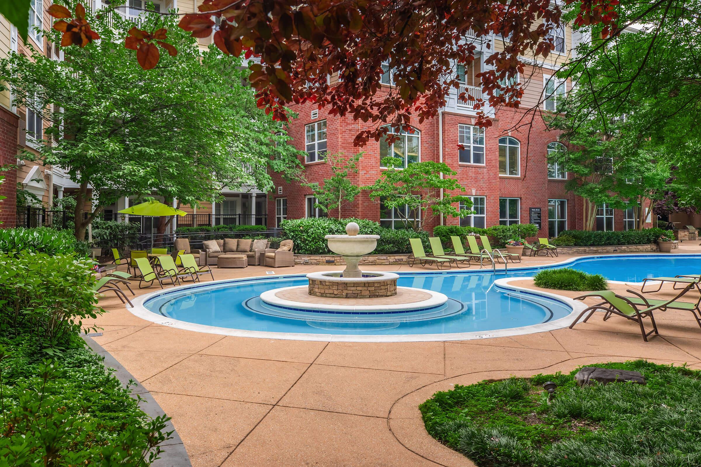 A serene pool area featuring a circular fountain at its center, surrounded by light-colored lounge chairs and lush greenery. The backdrop includes brick buildings with large windows, shaded by trees, creating a relaxing outdoor atmosphere.