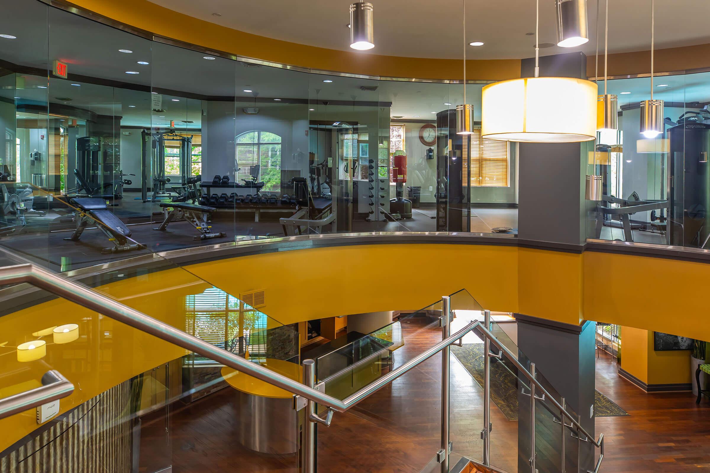 A modern gym interior featuring a spacious workout area with various exercise equipment. The design includes large windows allowing natural light, warm wood floors, and sleek, contemporary decor. A staircase with a curved railing connects different levels, enhancing the open and inviting atmosphere.