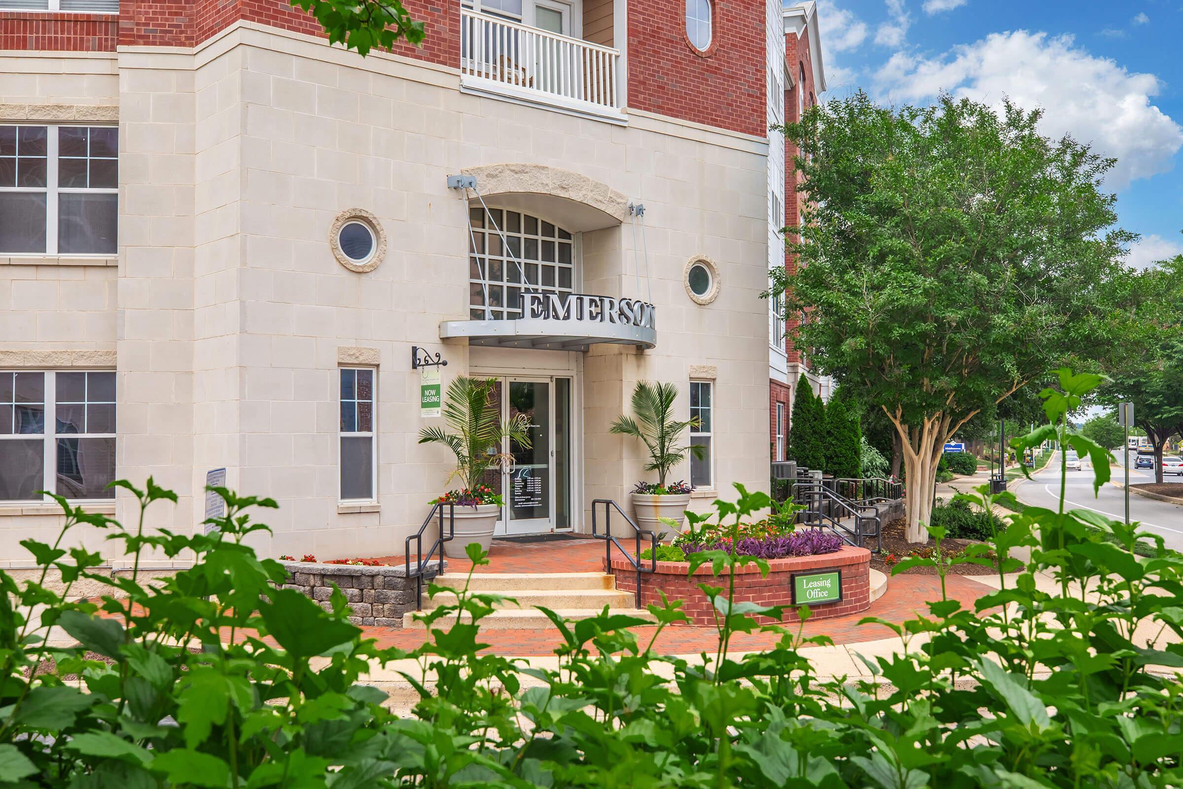 Facade of the Emerson residential building featuring a welcoming entrance with large glass doors, lush landscaping in the foreground, and a clear blue sky in the background. The building showcases a blend of brick and stone materials with a modern design.