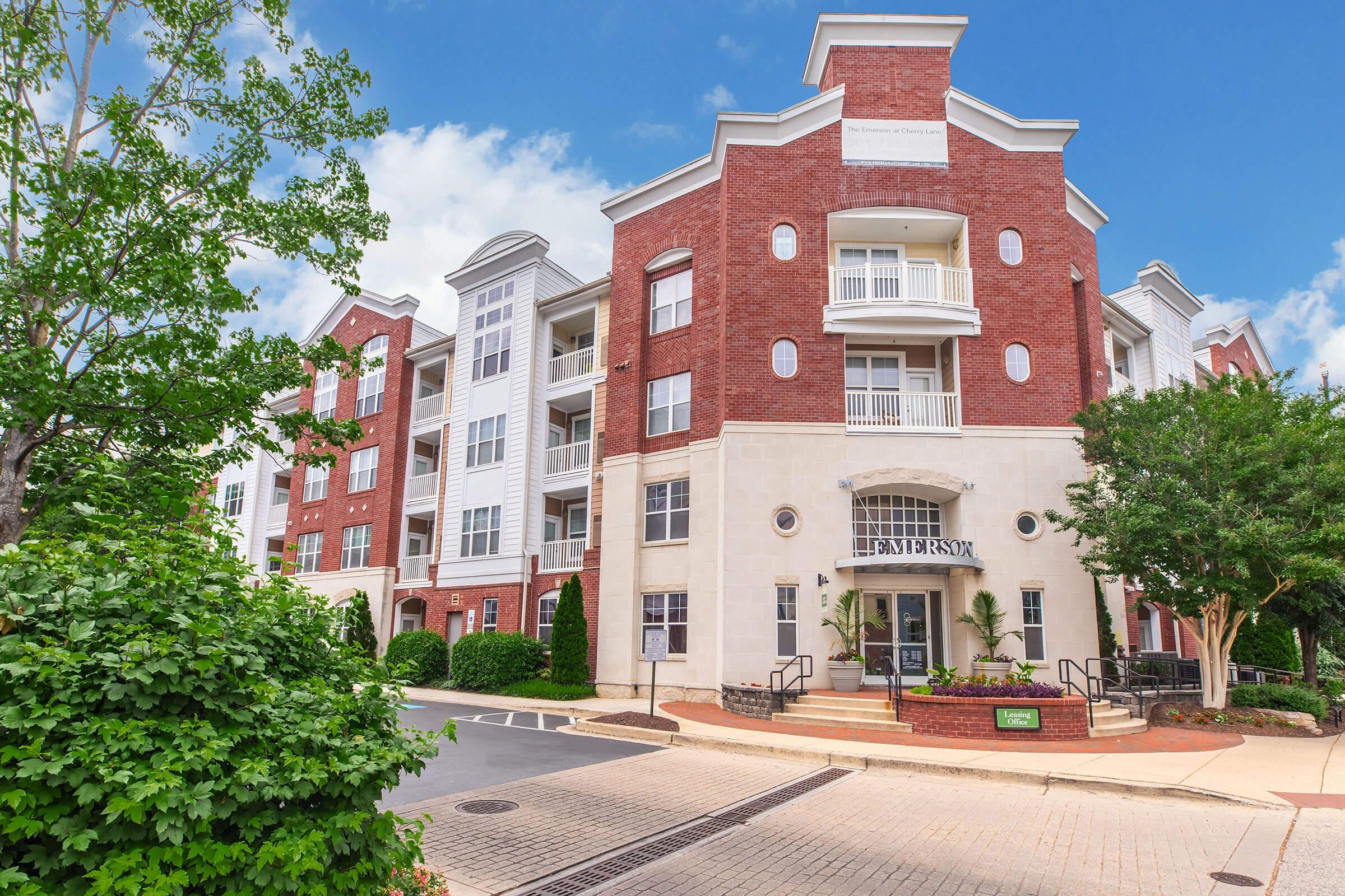 A modern brick apartment building with multiple stories, featuring large windows and balconies. The entrance is highlighted by steps and landscaped greenery. The sky is bright and partly cloudy, enhancing the building's contemporary aesthetic.