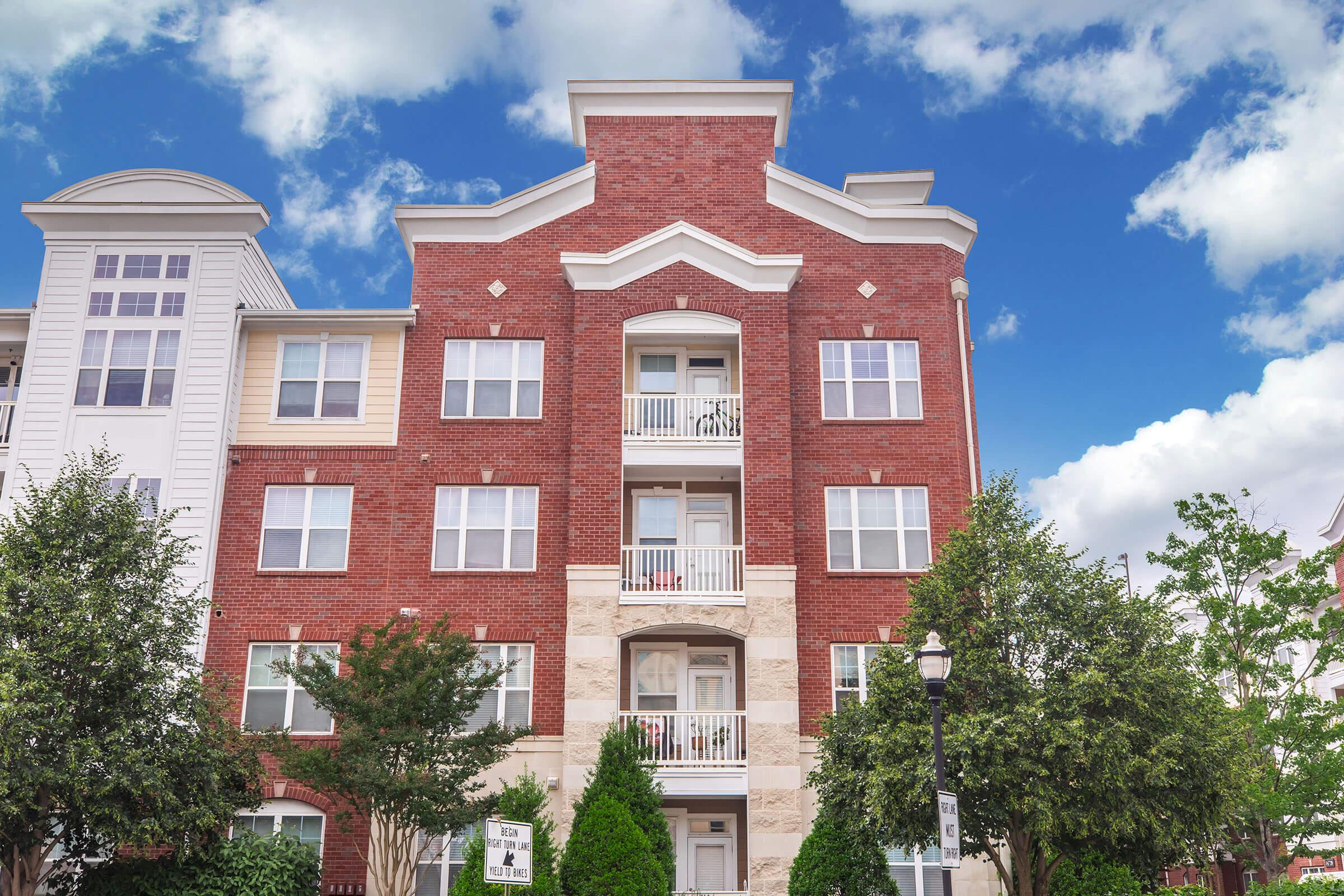 A red-brick apartment building with multiple floors and balconies, surrounded by green trees. The sky is partly cloudy, adding a bright atmosphere to the scene. The building features a mix of architectural styles, including large windows and decorative elements on the façade.