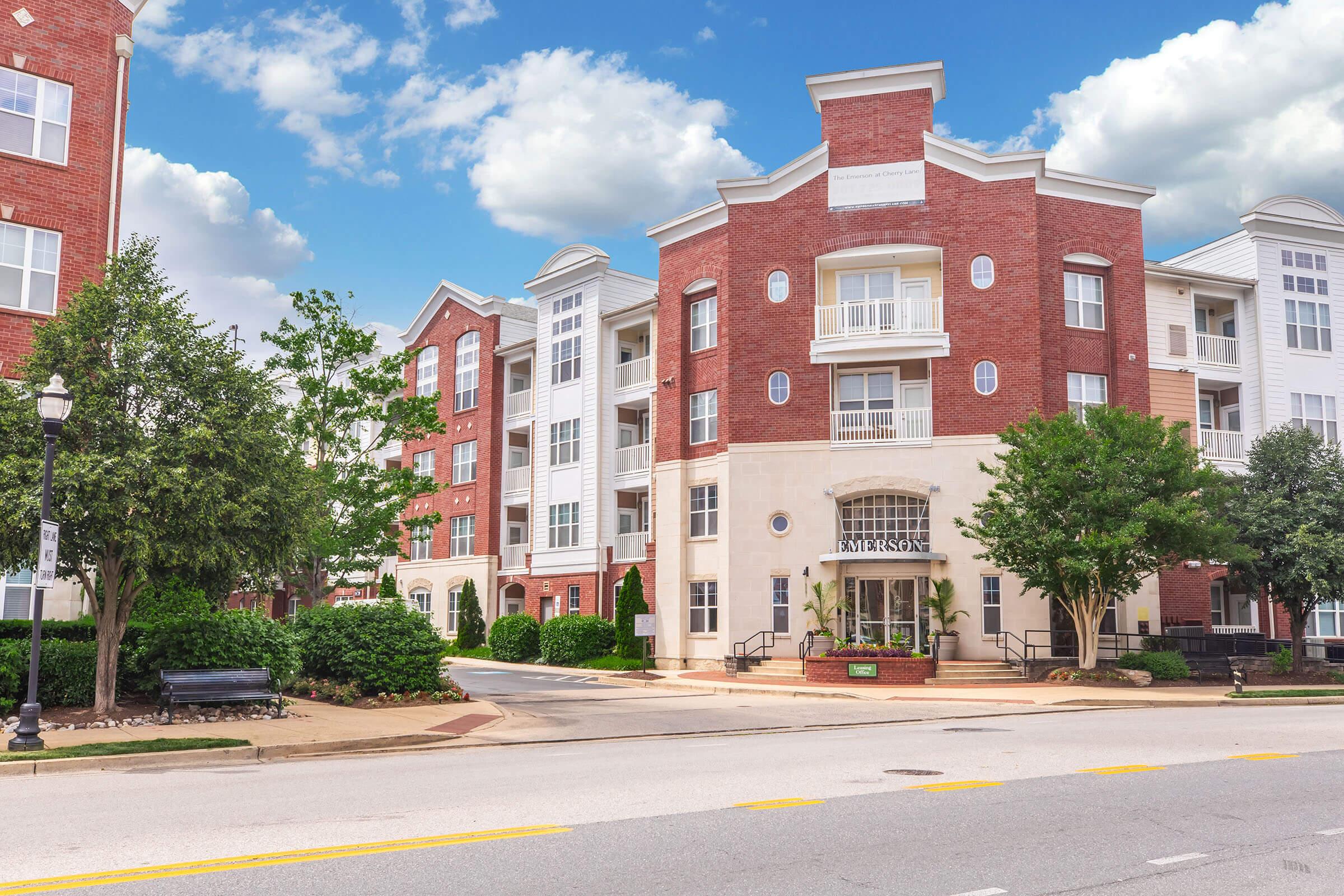 A well-maintained modern multi-story apartment complex featuring red brick architecture. The entrance is adorned with lush greenery and small trees. A wide pathway leads to the main doors, while the sky above is partly cloudy, creating a pleasant urban atmosphere.