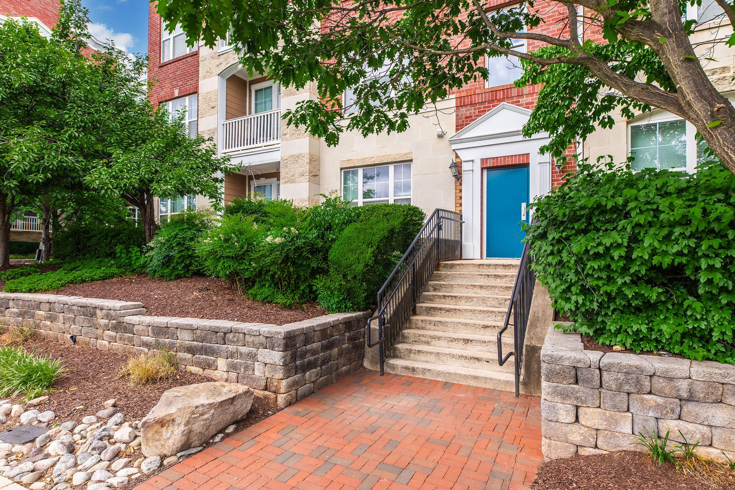 A landscaped outdoor entrance to a modern building featuring a set of stairs leading to a blue door. The area is surrounded by lush greenery, including shrubs and trees, with a stone and brick pathway leading up to the steps. The sky is partly cloudy, adding a bright, inviting atmosphere.