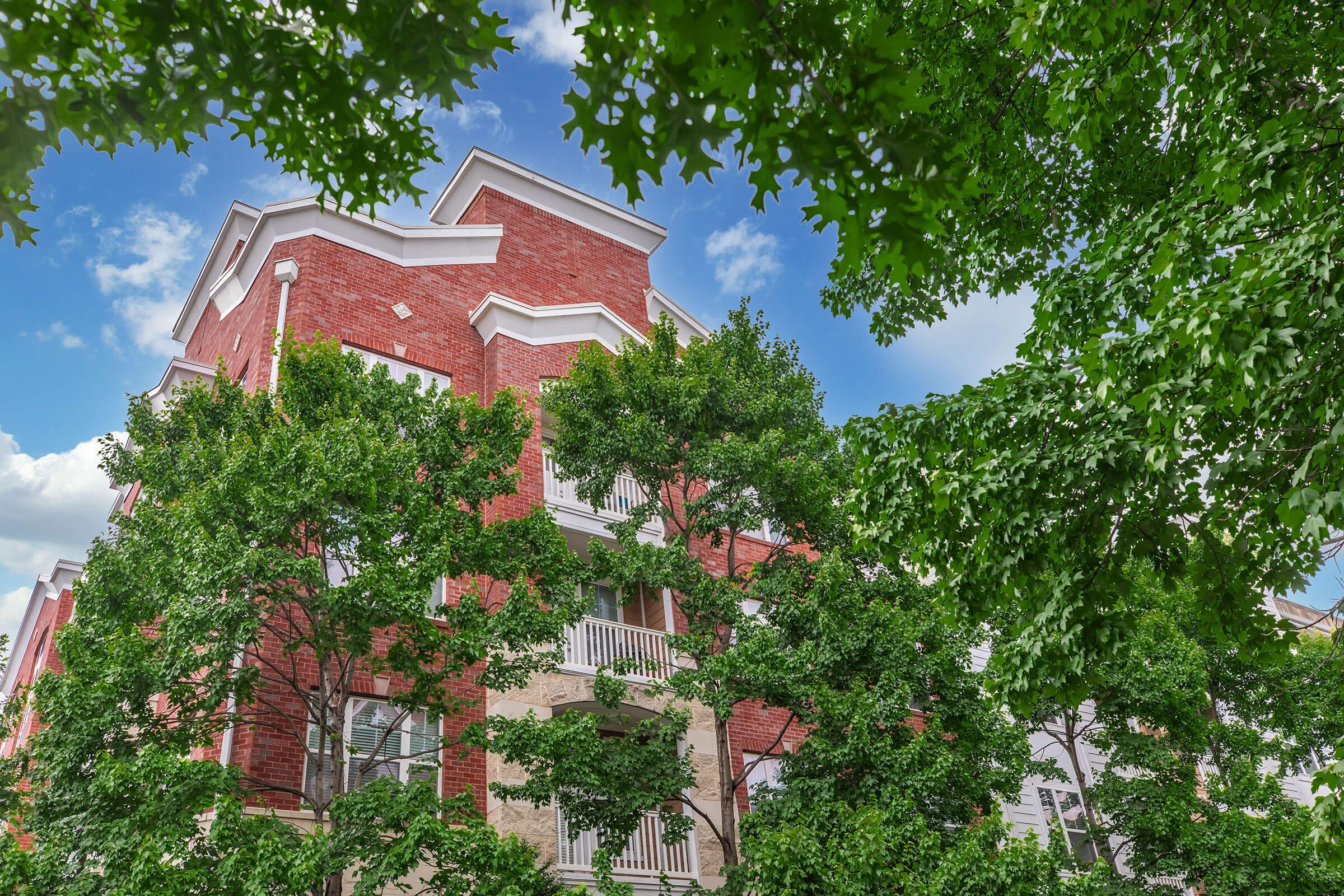 A red brick building partially obscured by lush green trees, under a bright blue sky with scattered clouds. The architecture features multiple levels and balconies, creating a vibrant urban scene that blends nature with city life.