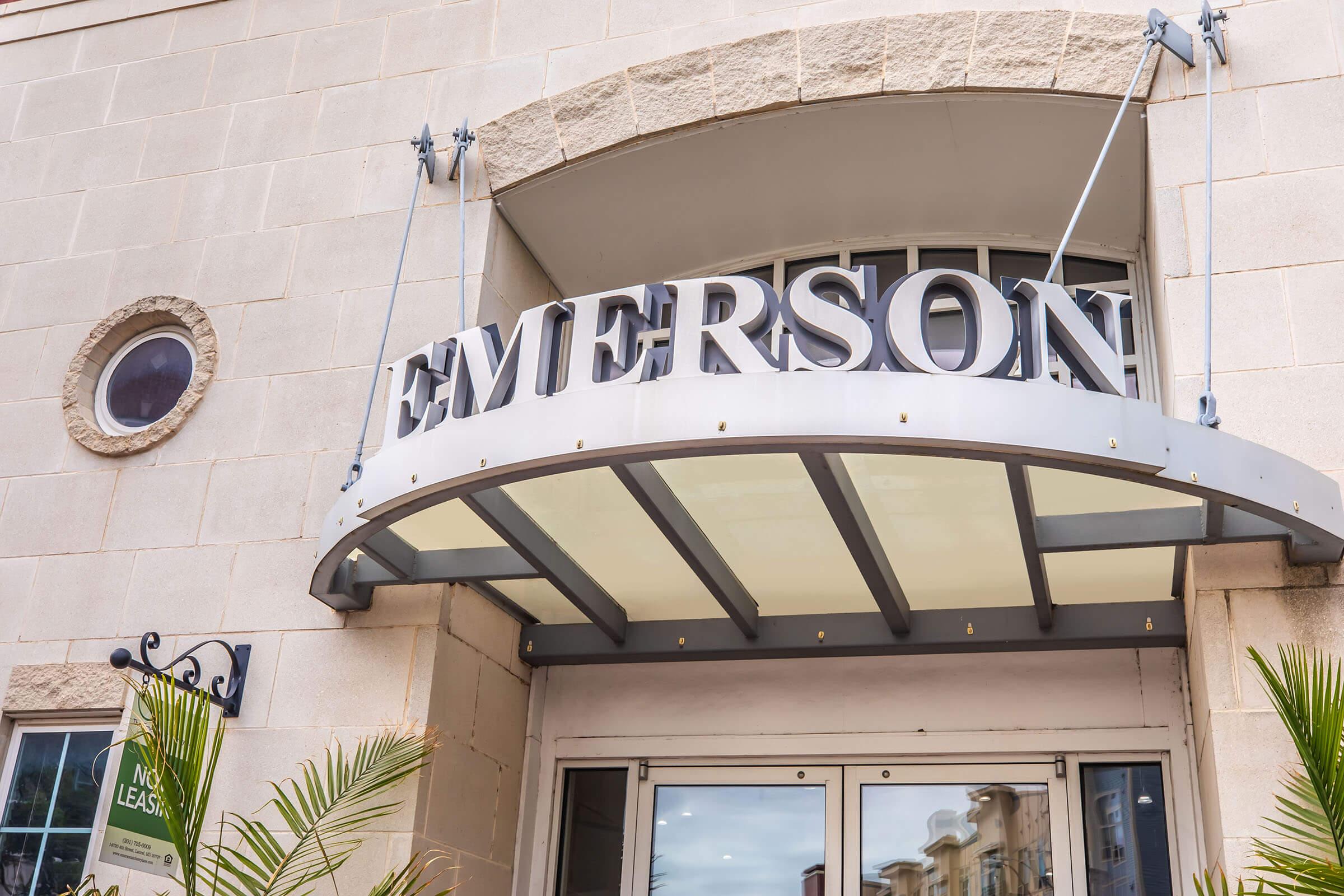 Signage for "EMERSON" displayed prominently above an entrance, with a modern awning. The facade is made of light-colored stone, and there is a decorative circular window nearby. Greenery is visible at the bottom, suggesting a welcoming entrance.