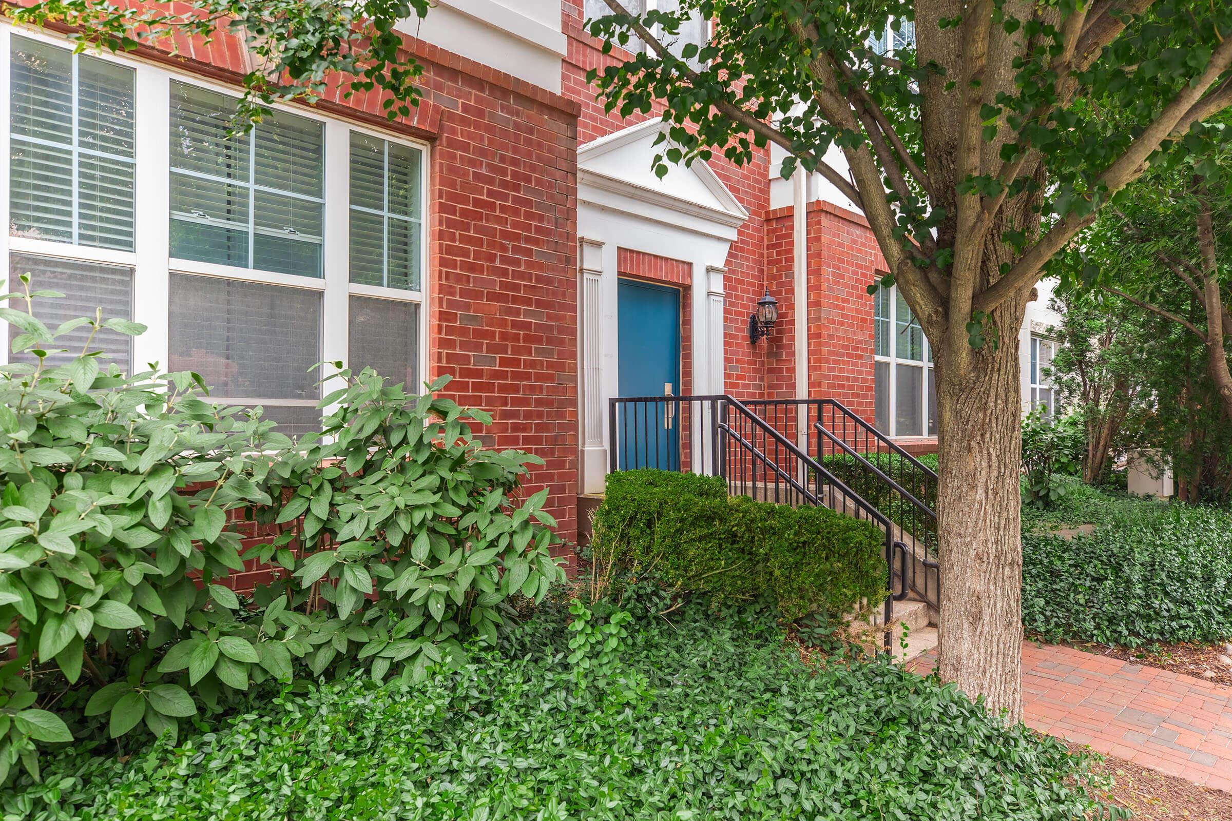 A brick building with a blue front door. There are large windows beside the door, framed by green bushes and ivy on the ground. A small set of stairs leads up to the entrance, and a tree is visible on the right side of the image, adding to the greenery surrounding the area.