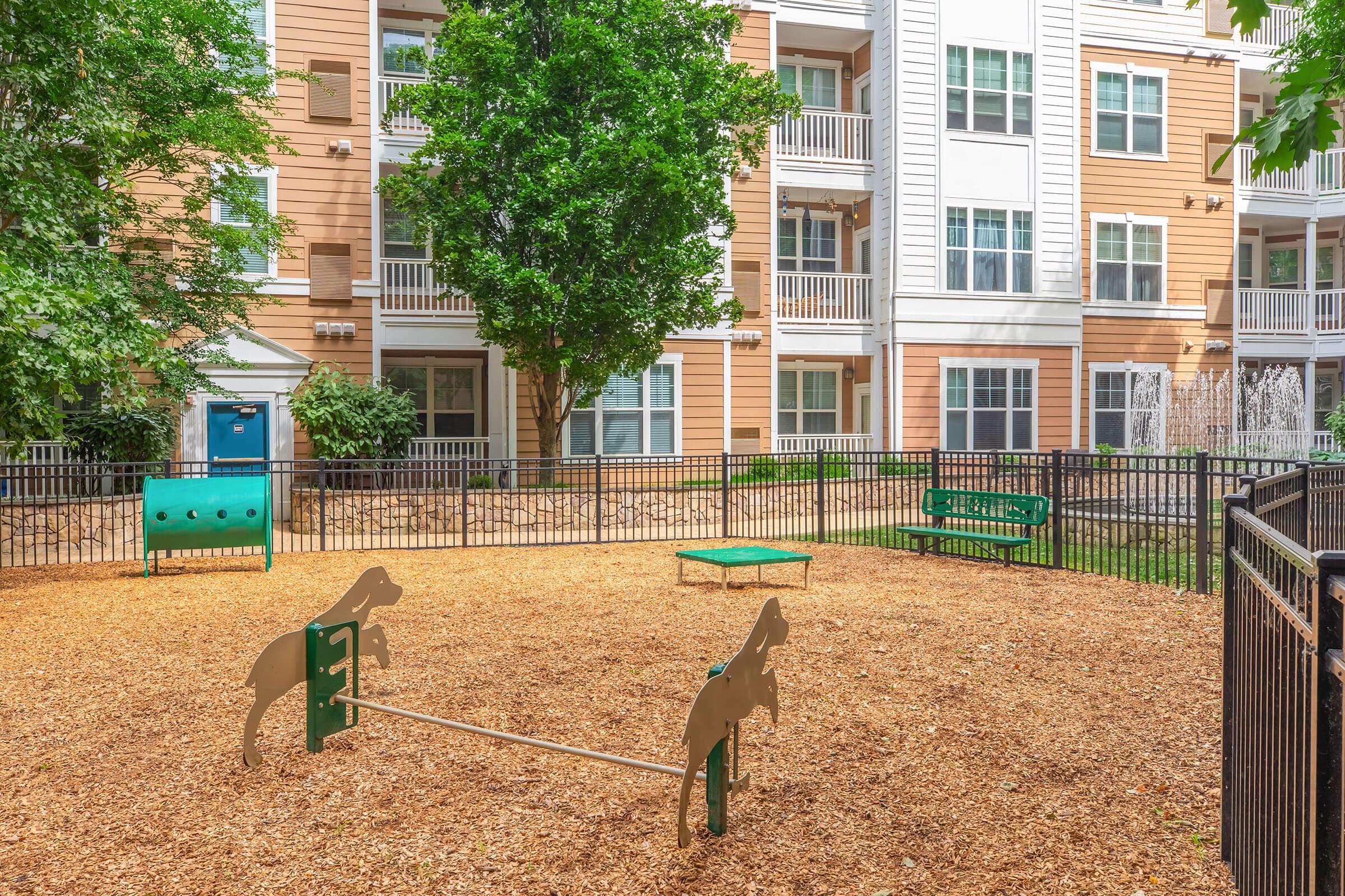 A dog park featuring a fenced area with wood chips, a small green agility frame, and colorful dog-inspired structures. Surrounding it are apartment buildings with balconies and greenery, providing a pet-friendly space for residents.