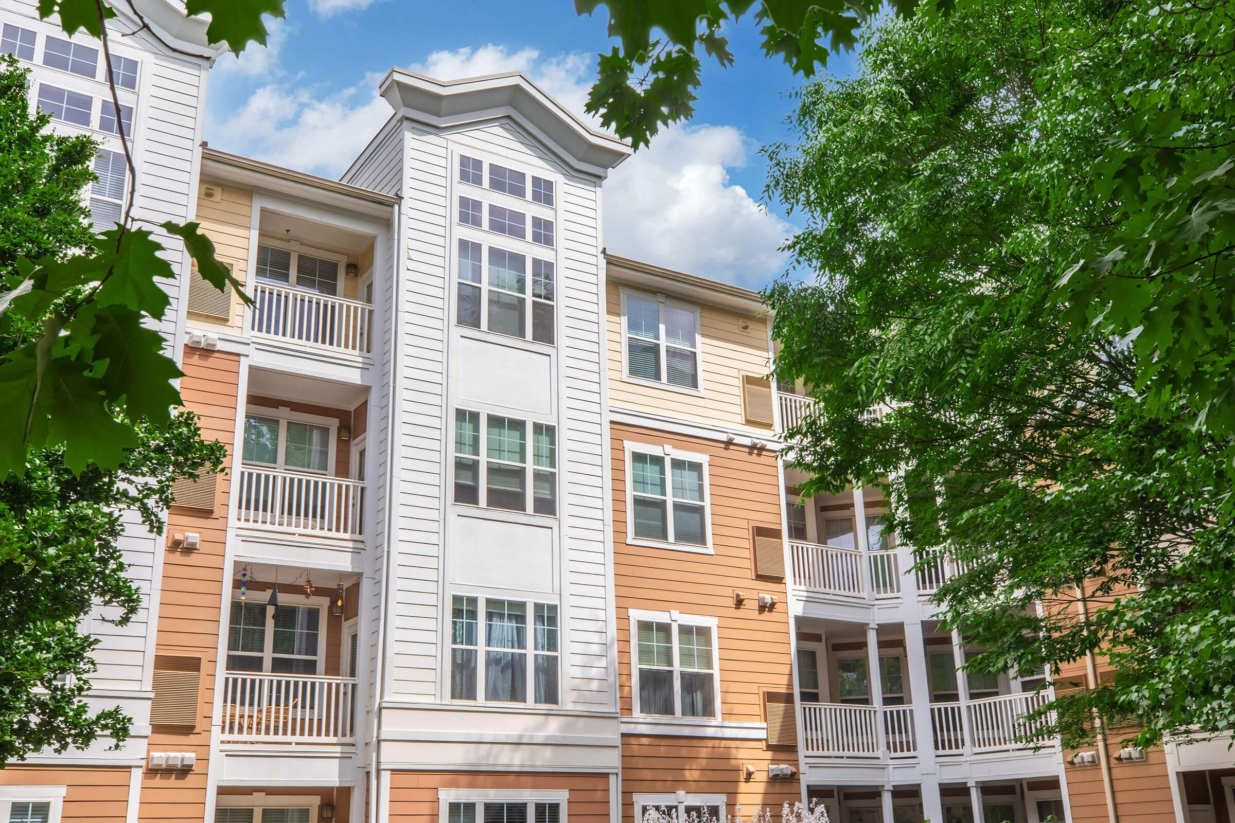 A multi-story apartment building with a mix of orange and white siding, featuring numerous windows and balconies. Lush green trees frame the scene, and the sky is partly cloudy, creating a bright and inviting atmosphere.
