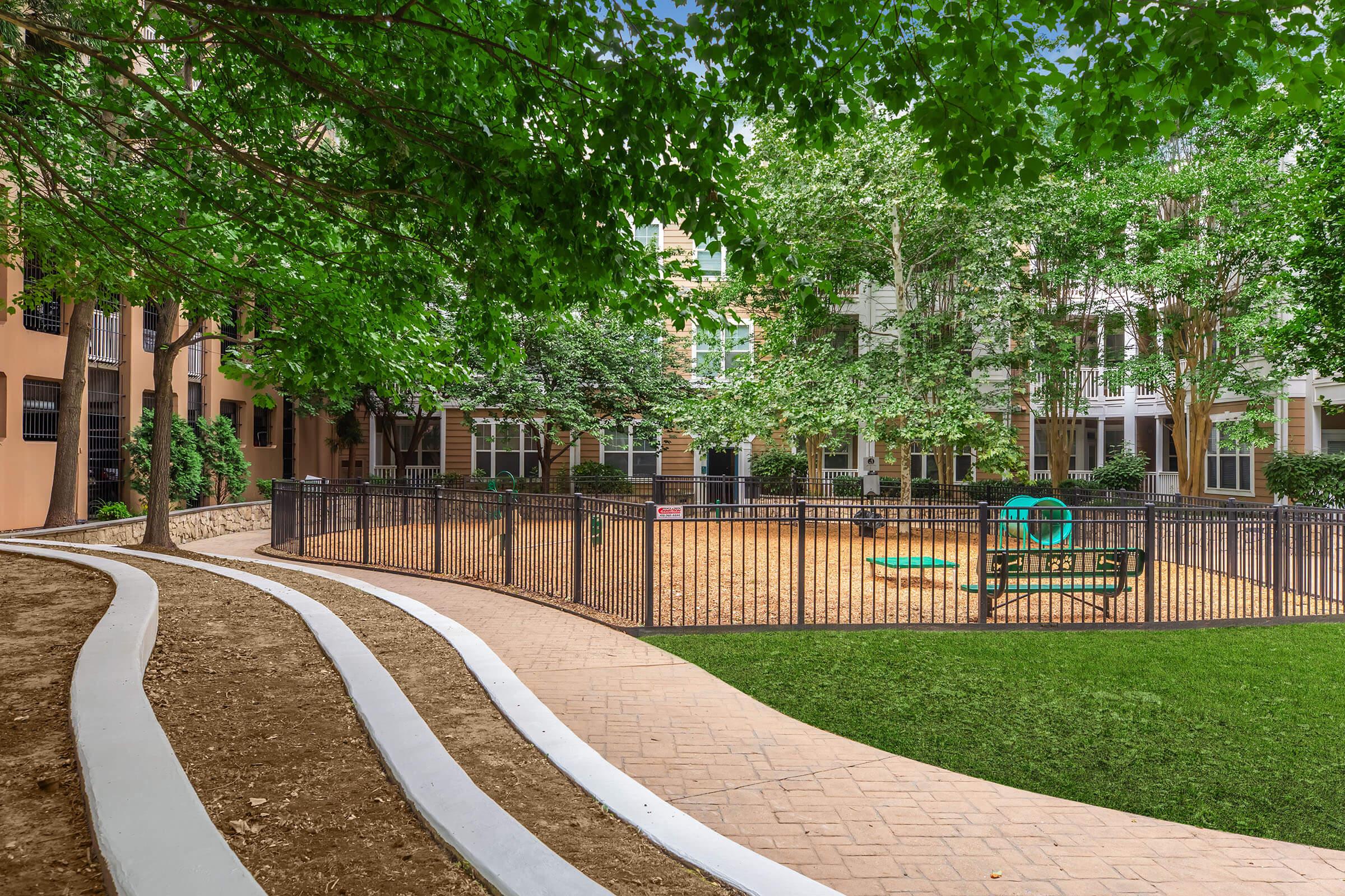 A green, landscaped courtyard featuring a sandy play area surrounded by a black fence. There are trees providing shade, and walking paths made of concrete and gravel leading towards the play area. The scene is peaceful and inviting, ideal for relaxation and outdoor activities.