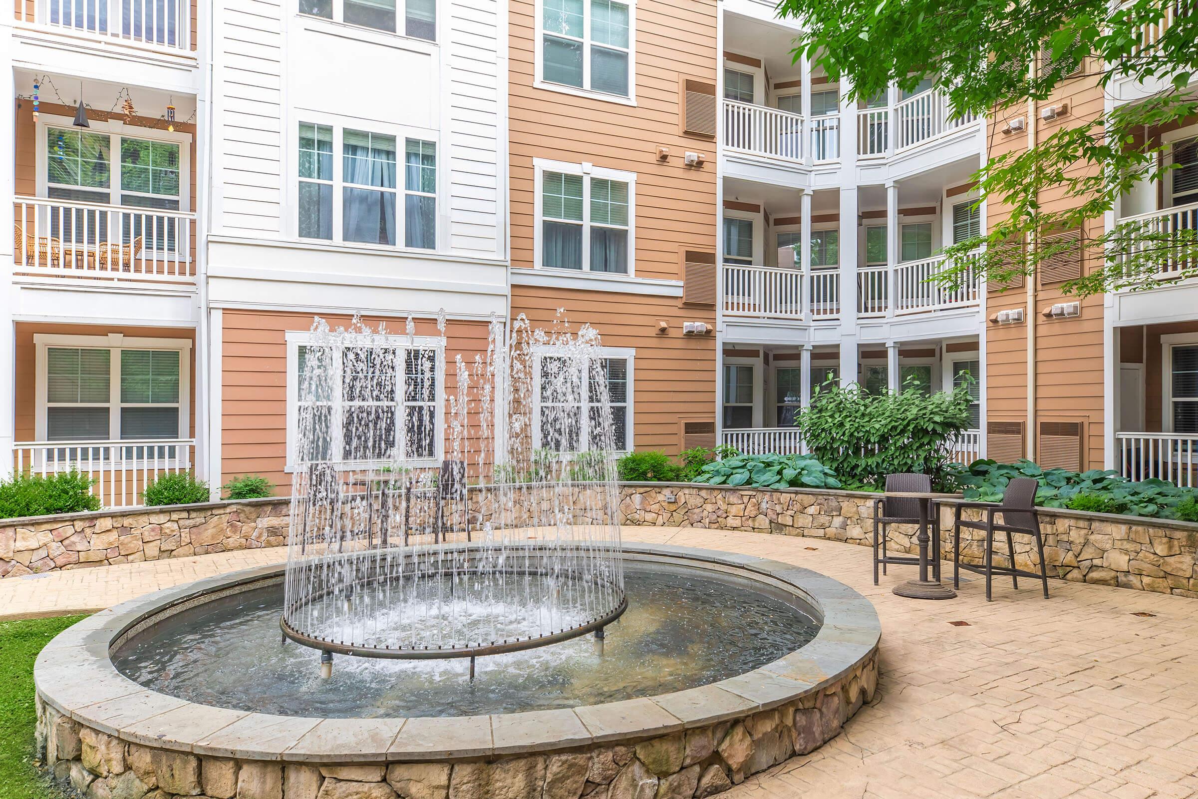 A tranquil courtyard featuring a circular stone fountain with water cascading into a round pool. Surrounding the fountain are lush green plants and a paved walkway. In the background, there are multiple modern apartment building balconies with white railings. Two chairs and a small table are set near the fountain.