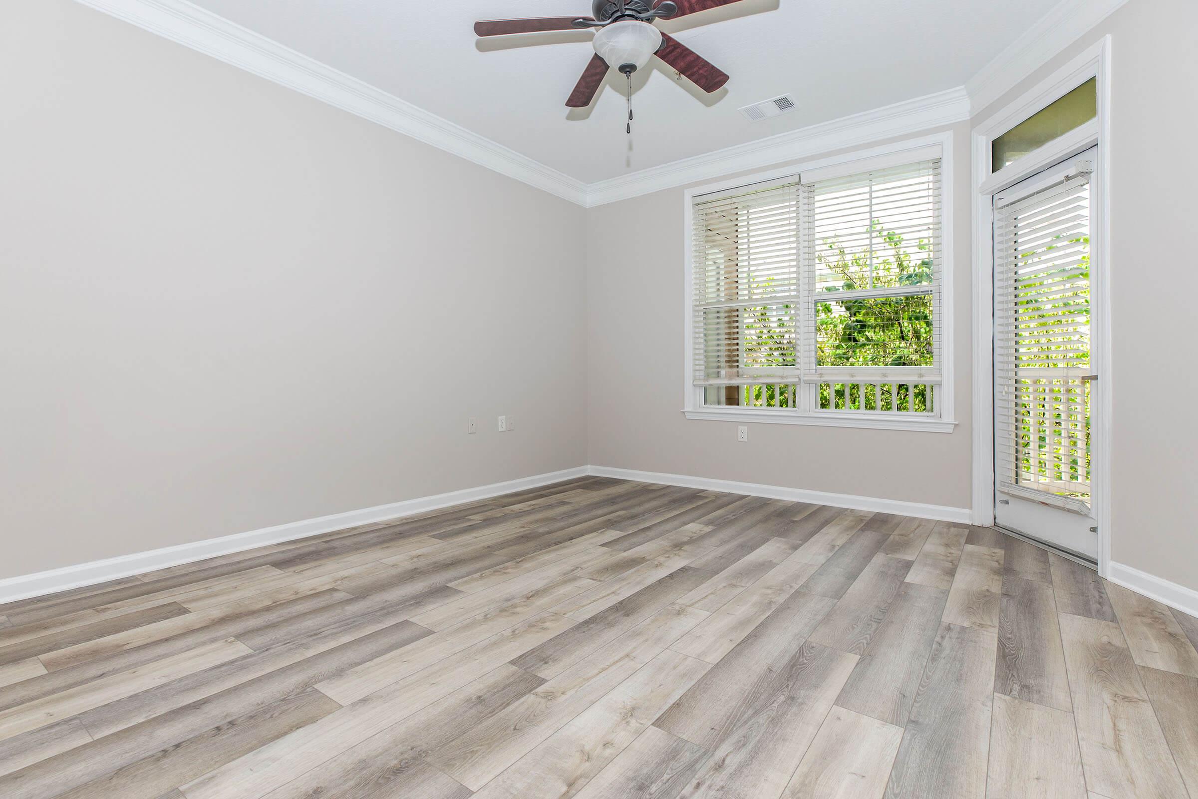 A spacious, minimally furnished room featuring light wood-look flooring, a ceiling fan, and large windows with blinds. The walls are painted a soft neutral color, enhancing the brightness of the space. A door leads to a balcony or outdoor area, surrounded by greenery.
