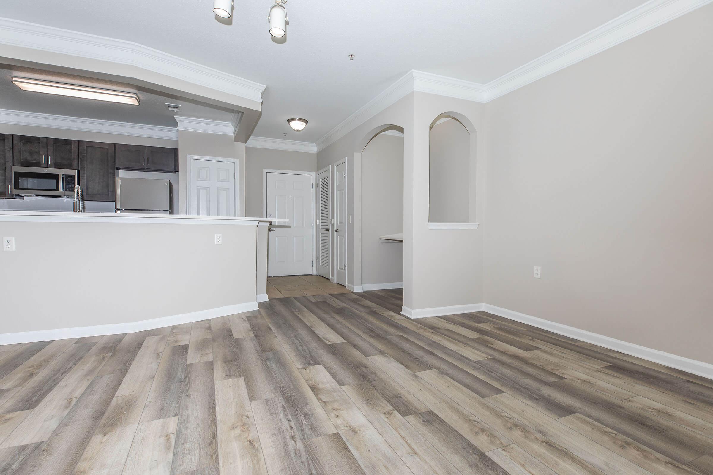 A spacious, modern living area with light-colored walls and wood-like flooring. To the left, there's a kitchen with dark cabinetry and stainless steel appliances. The room features crown molding and has an open layout with an arched entryway leading to another room. Natural light enters the space, creating a welcoming atmosphere.