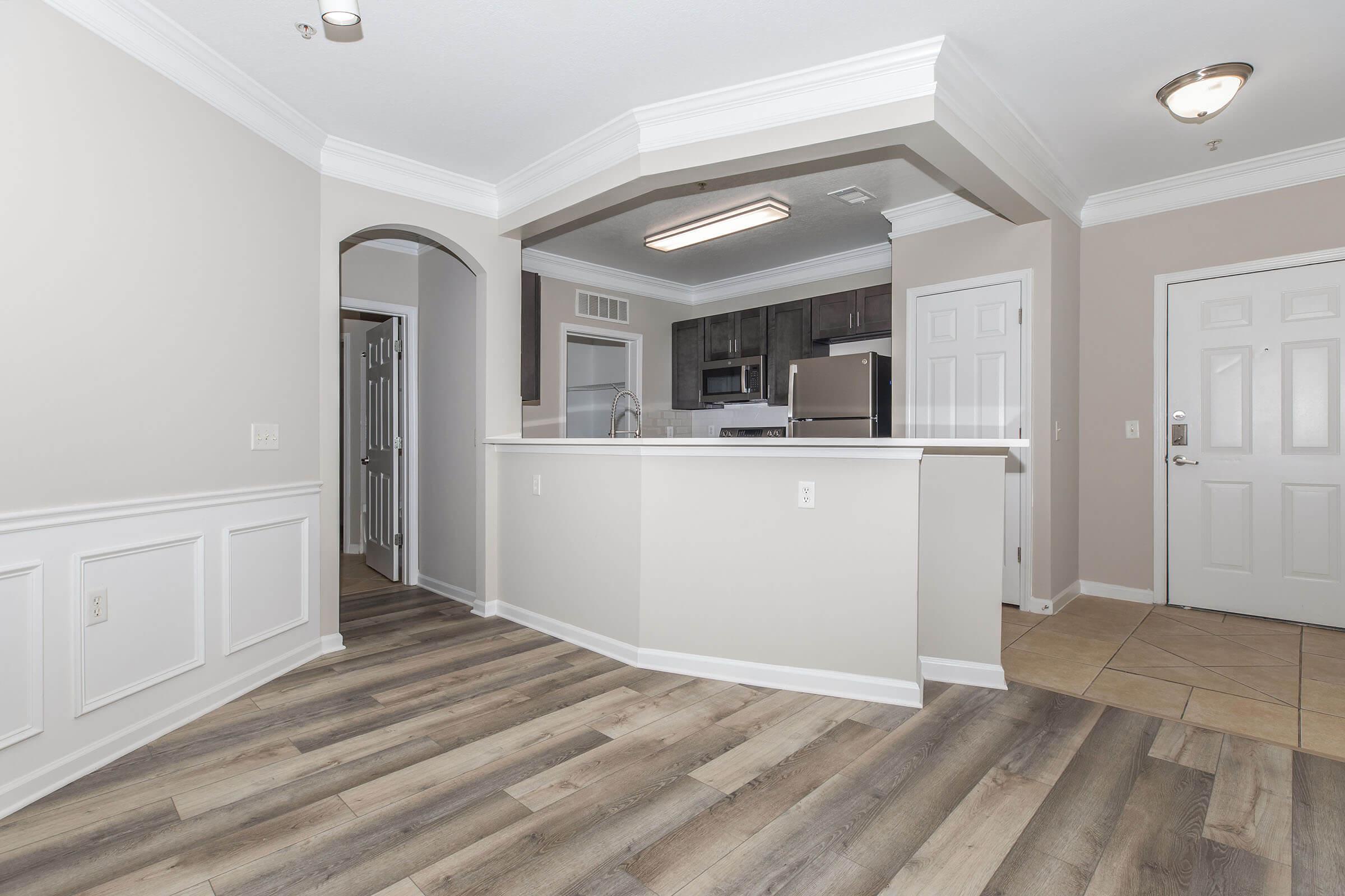 A view of a modern apartment interior featuring a spacious layout. The room includes a kitchen area with dark cabinets and stainless steel appliances, a doorway leading to another room, and a front entrance. The flooring is a mix of wood-like planks and tile, with elegant wainscoting along the walls.