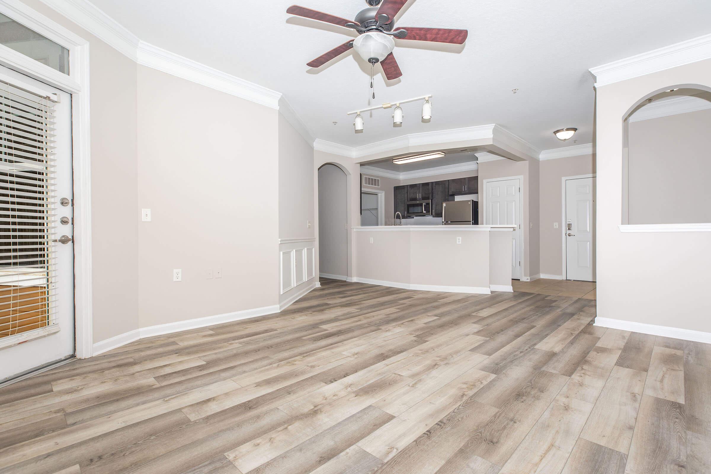 Spacious, light-filled living area with modern wood-style flooring, ceiling fan, and archways. Open floor plan leading to a kitchen area with dark cabinetry. Sliding glass doors open to an outdoor space, enhancing the inviting atmosphere. Neutral-colored walls create a warm and welcoming environment.