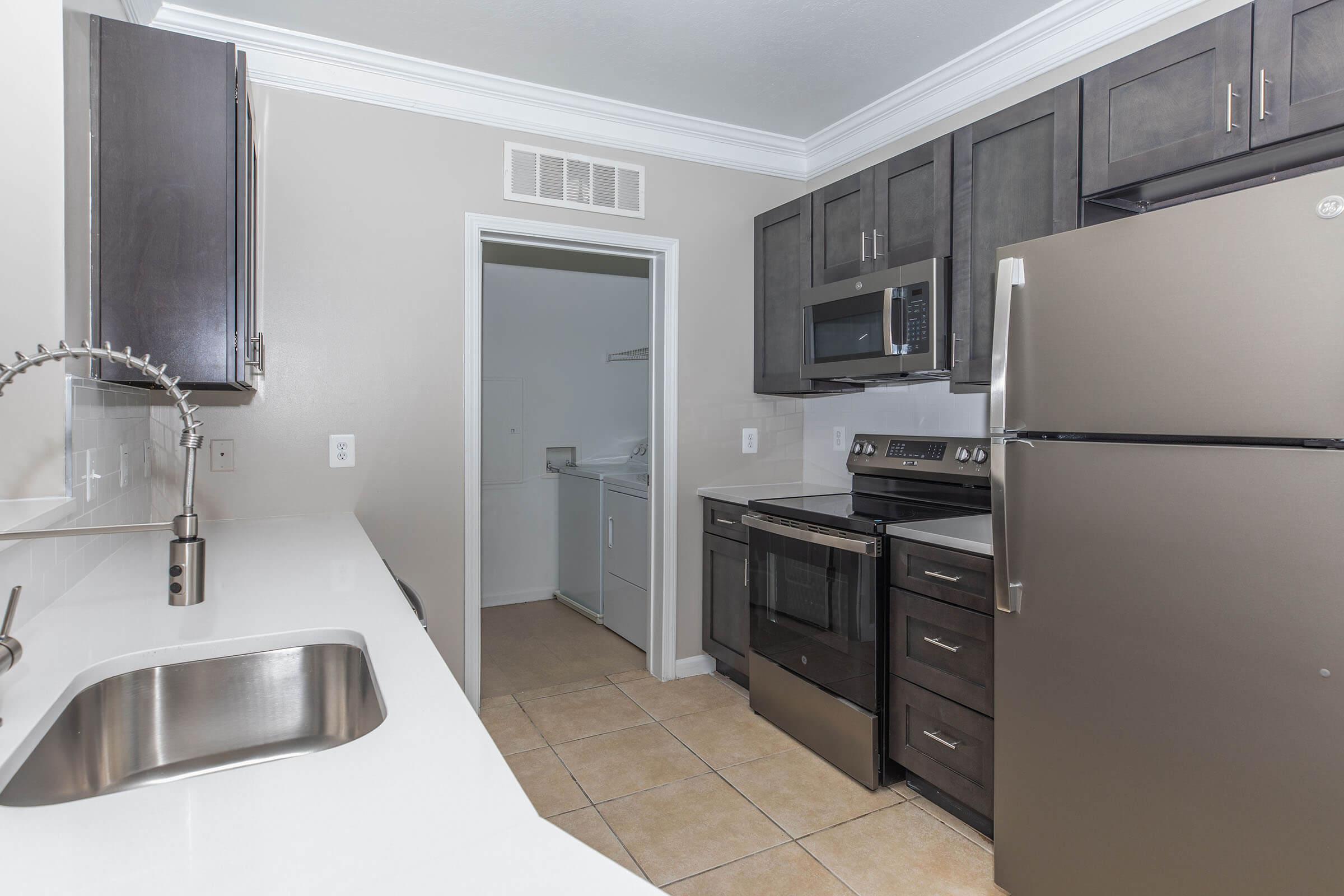 Modern kitchen featuring dark cabinetry, stainless steel appliances including a refrigerator, oven, and microwave. A white countertop with a sink is visible, along with beige tile flooring. A doorway leads to a laundry area. The kitchen has a clean, contemporary design with neutral colors.