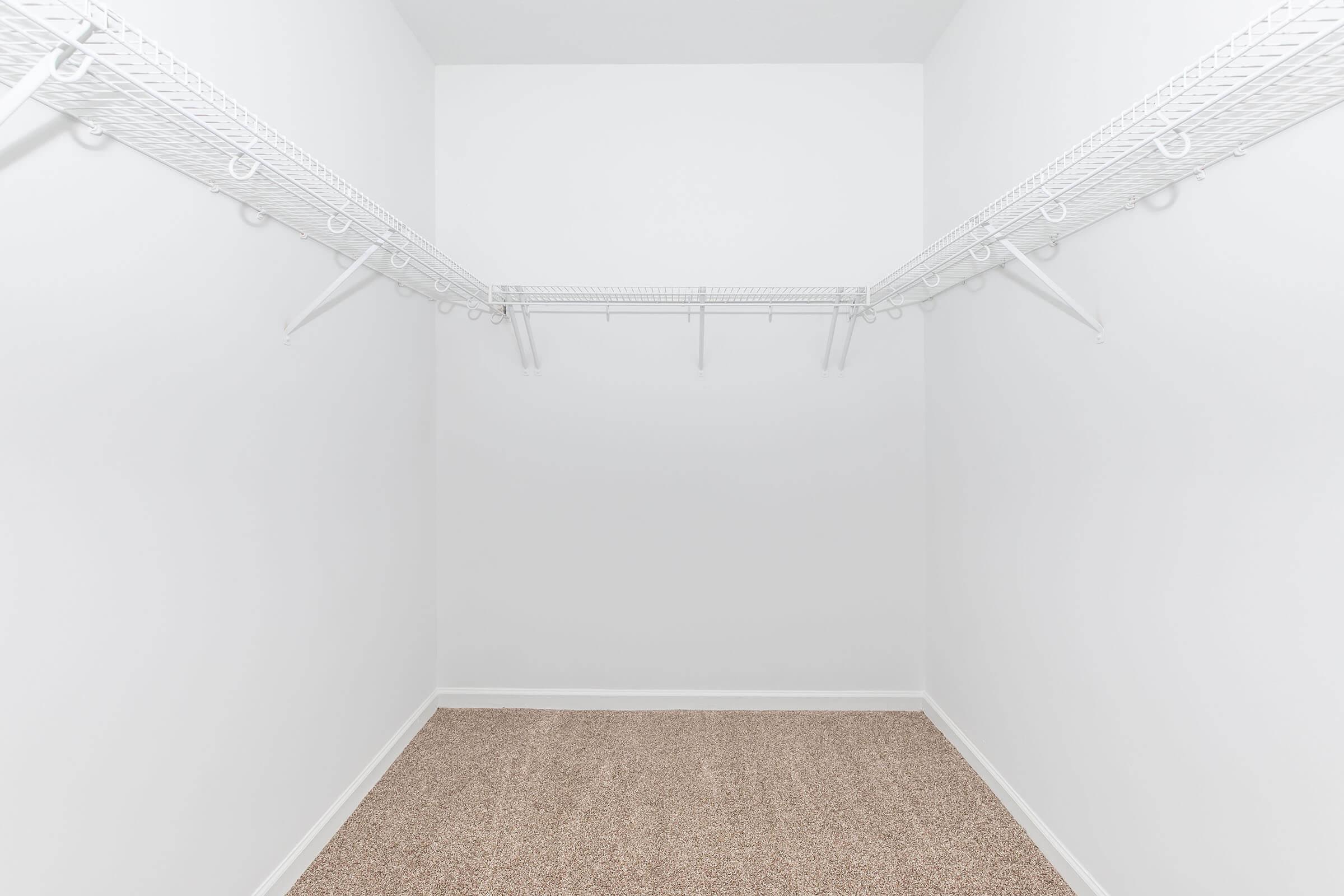 Empty closet with white walls and a beige carpeted floor. The closet features a white wire shelving unit along one wall, with several hooks for hanging items. The space is well-lit and has no visible items or decorations.