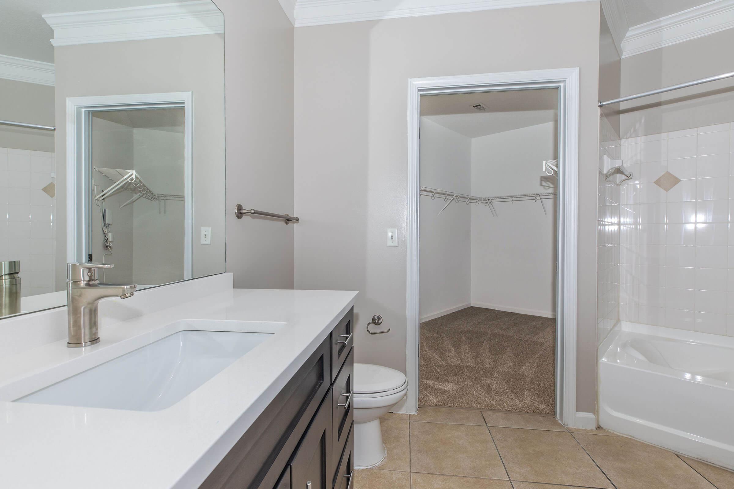 A modern bathroom featuring a light-colored tile floor, a dual-sink vanity with a white countertop, a toilet, and a large mirror. On one side, there is a door leading to a spacious walk-in closet with shelving, and on the other side, a bathtub with a shower fixture. The walls are painted a soft beige.