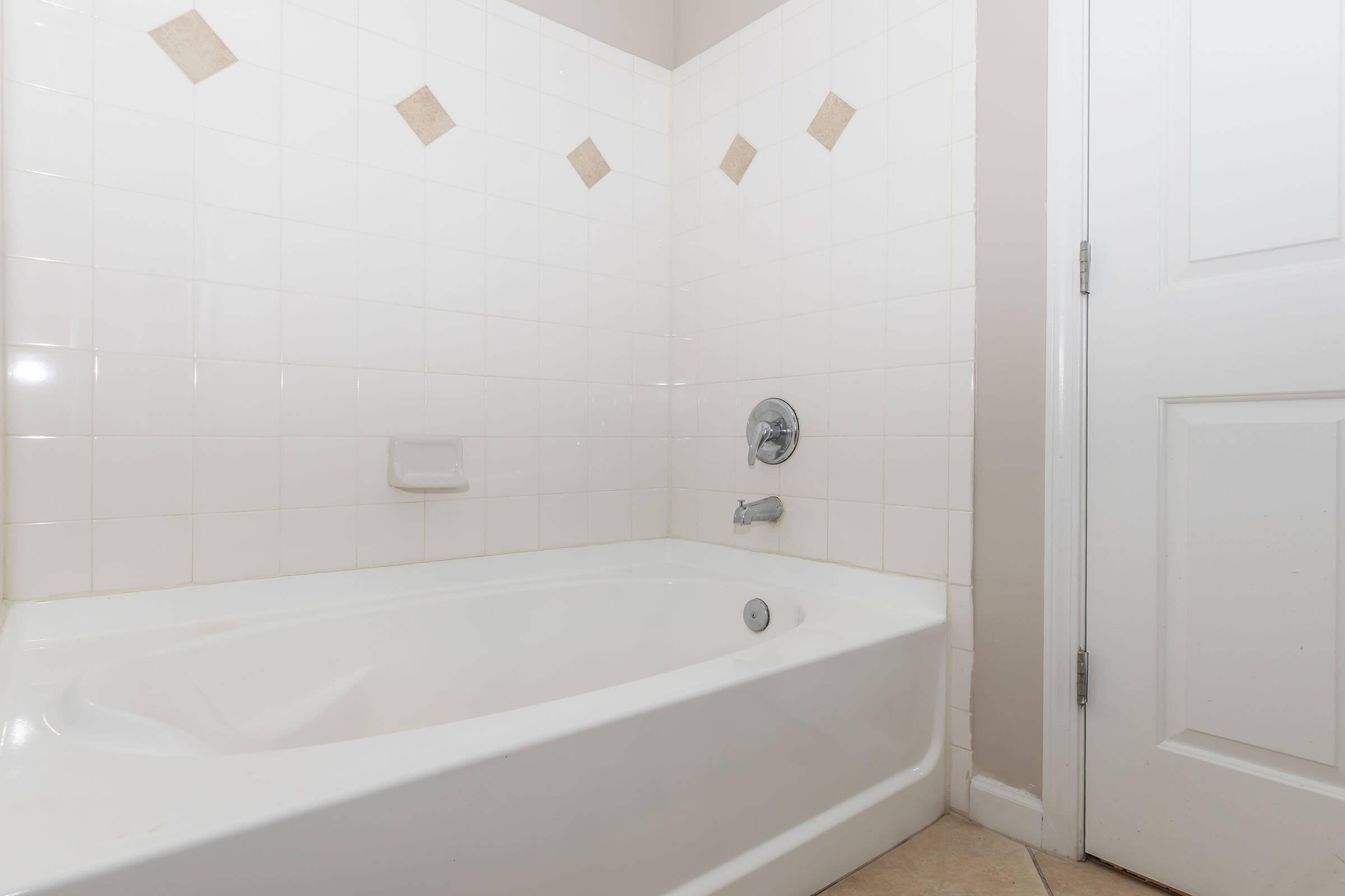 A clean and simple bathroom setup featuring a white bathtub with a chrome faucet and a showerhead. The walls are tiled in white with decorative beige diamond accents. A white door is visible on the right, completing the minimalist aesthetic of the space.