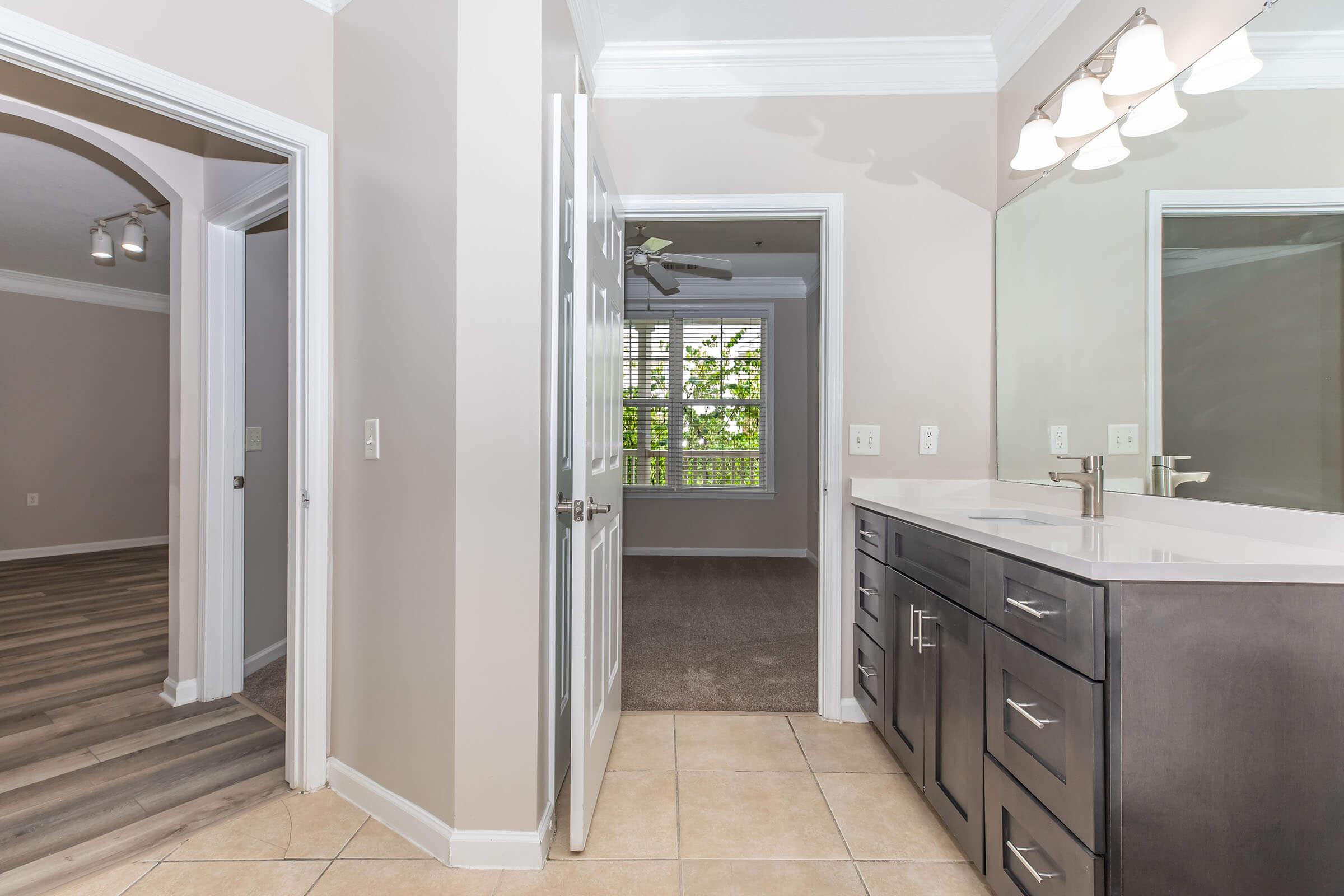 A modern bathroom with a double vanity featuring dark cabinetry and a large mirror. The room has tiled flooring, and a doorway leads to a bright bedroom with a window overlooking greenery. Another doorway opens to a hallway with wood-like flooring and recessed lighting.