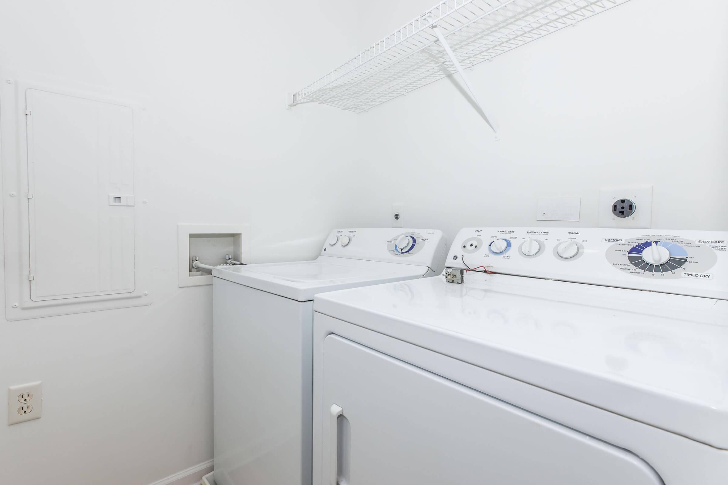 A clean, bright laundry room featuring a white washing machine and dryer side by side. There's a wire shelf above for storage, and a wall outlet and a small panel on the left side. The walls are painted white, creating a tidy and organized space for laundry tasks.