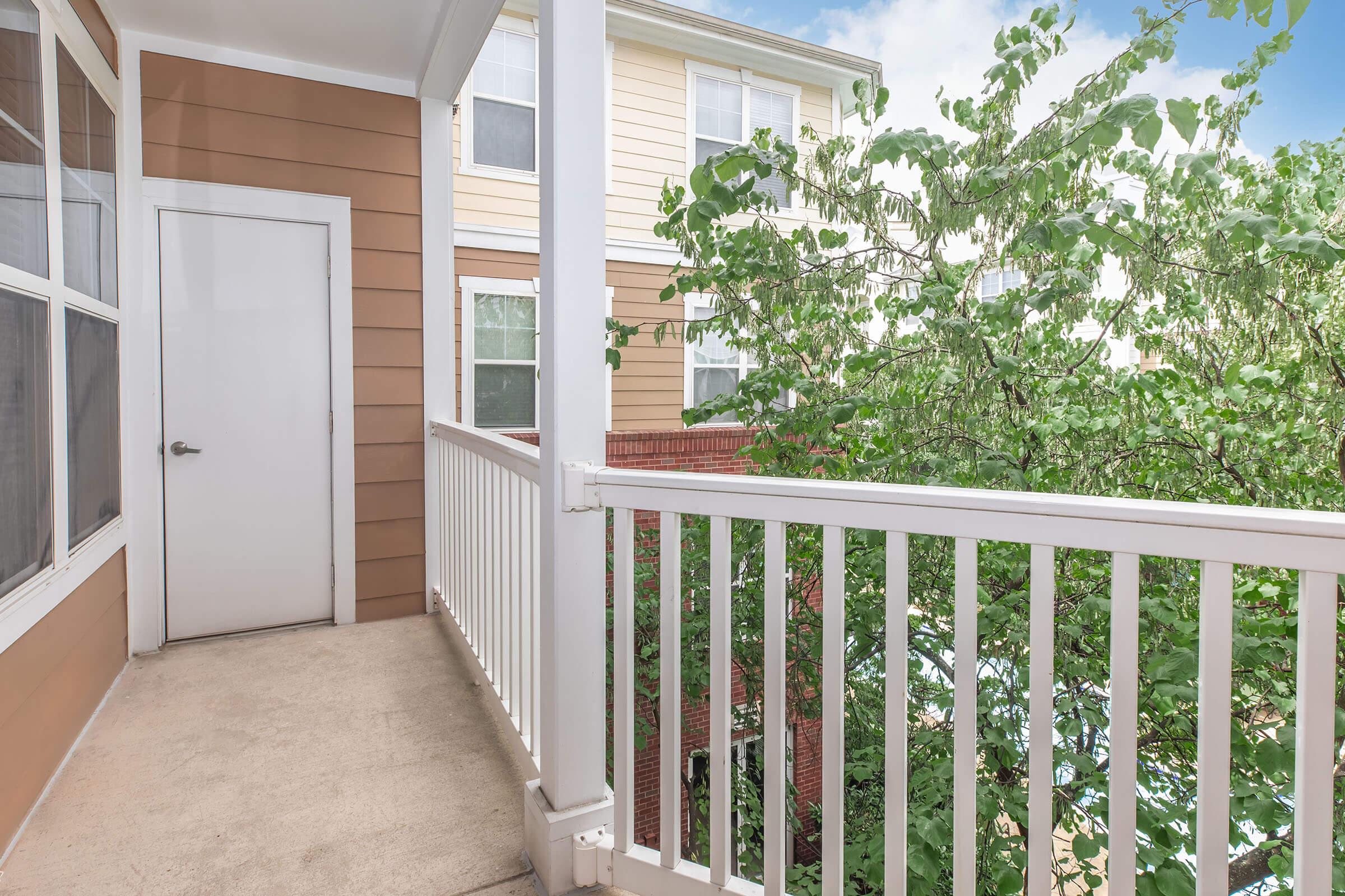 A view of a small balcony with white railing, featuring a door leading into a building. The balcony overlooks a green tree and adjacent buildings, with a bright sky in the background. The area is well-lit, suggesting a pleasant outdoor space.