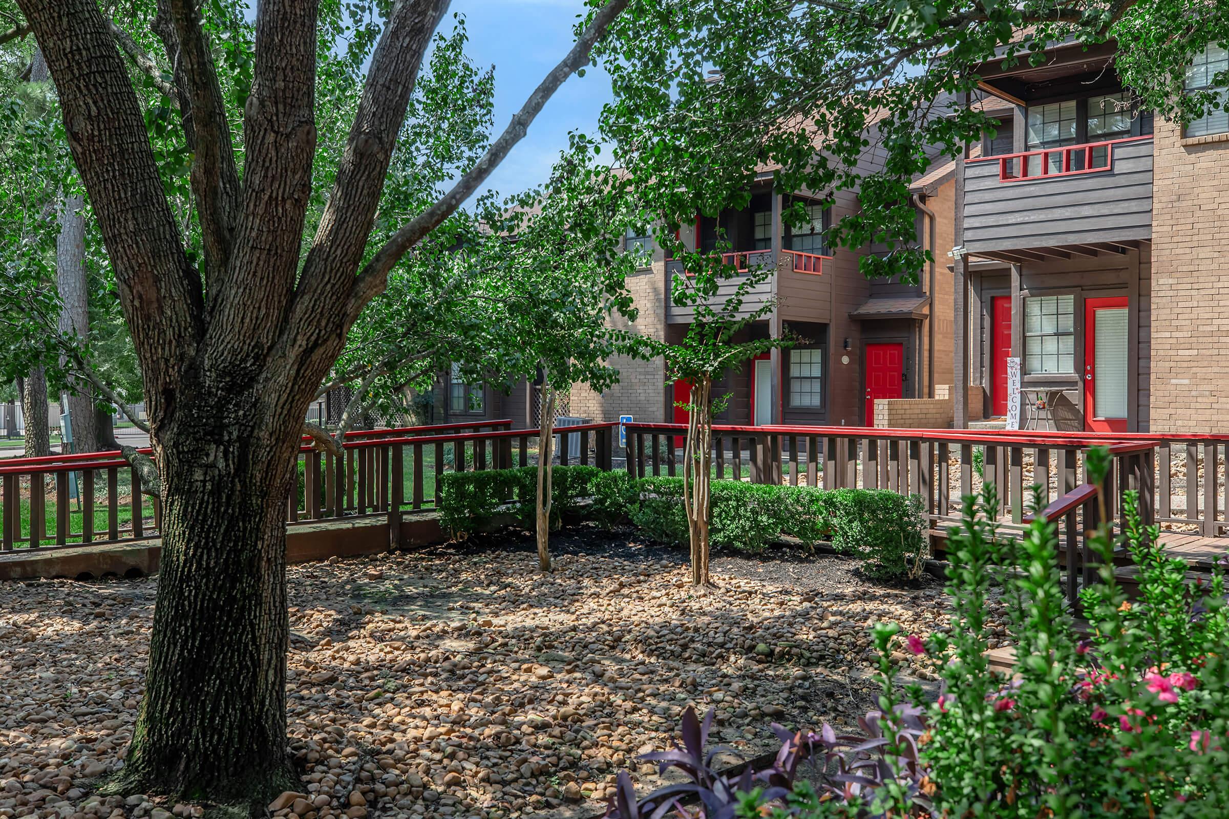 A landscaped courtyard featuring a large tree and decorative rocks, surrounded by brick buildings with red doors. Lush green plants and shrubs add color to the scene, creating a tranquil outdoor space for residents. Sunlight filters through the leaves, enhancing the inviting atmosphere.