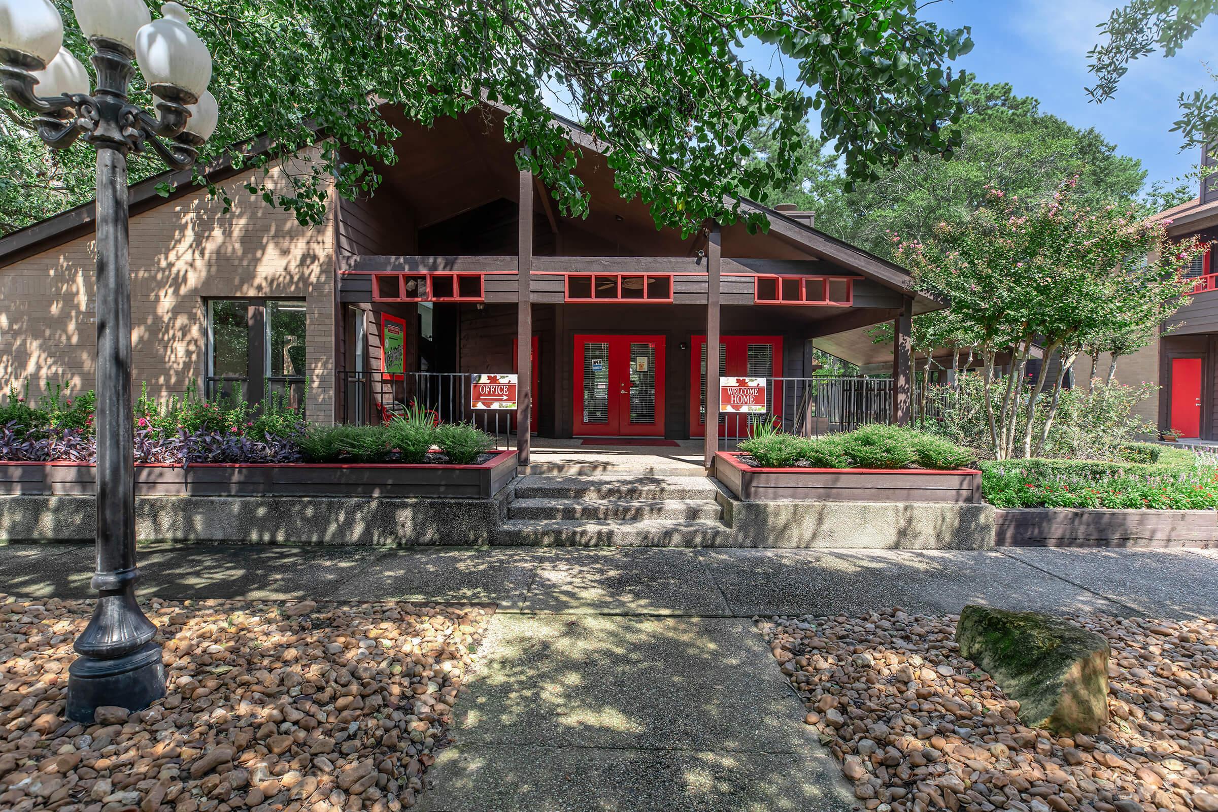 A welcoming lodge-style building with a sloped roof, featuring red accents and large front doors. The entrance is framed by greenery and flower beds, with a pathway made of stones leading up to it. Bright sunlight filters through the trees, creating a warm and inviting atmosphere.
