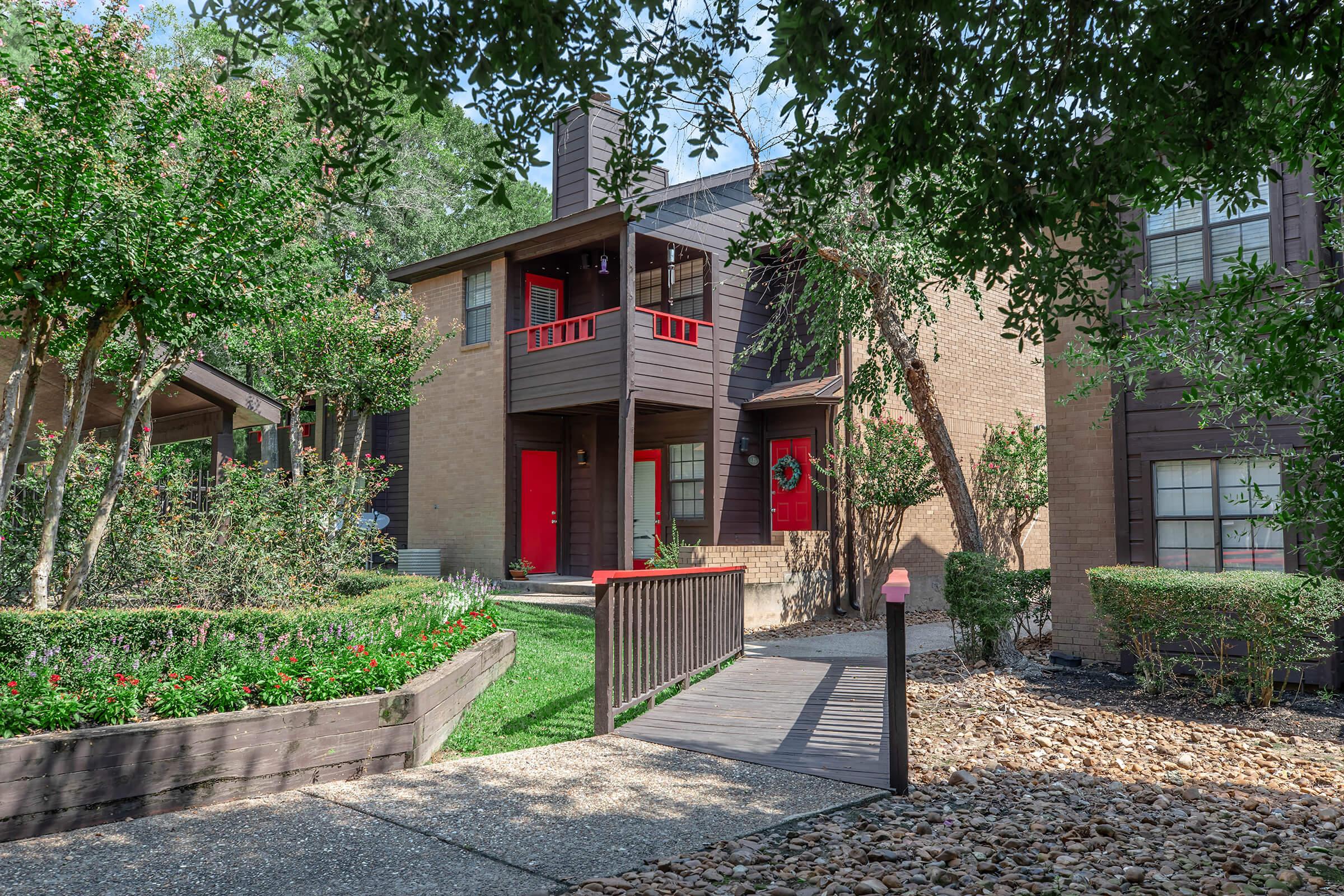 A peaceful residential scene featuring several brown brick apartment buildings with vibrant red doors. Lush greenery surrounds the entries, with well-maintained flower beds and trees providing shade. A wooden walkway leads to the main entrance, enhancing the inviting atmosphere of the complex.