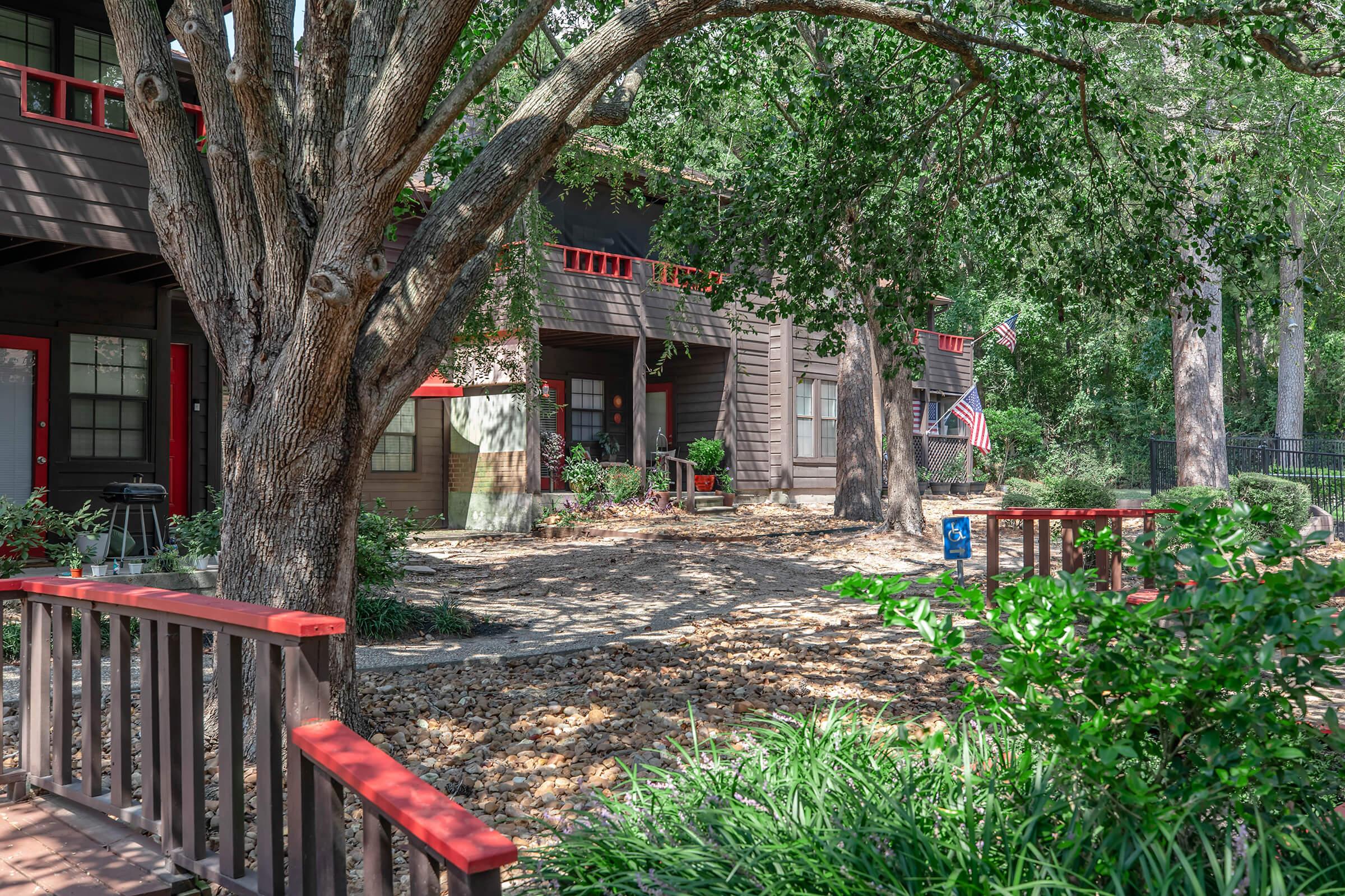 A cozy outdoor setting featuring a large tree with lush green leaves, red benches, and a pathway lined with light-colored stones. The background showcases a rustic wooden building with red accents and American flags. The scene is peaceful and inviting, surrounded by greenery.