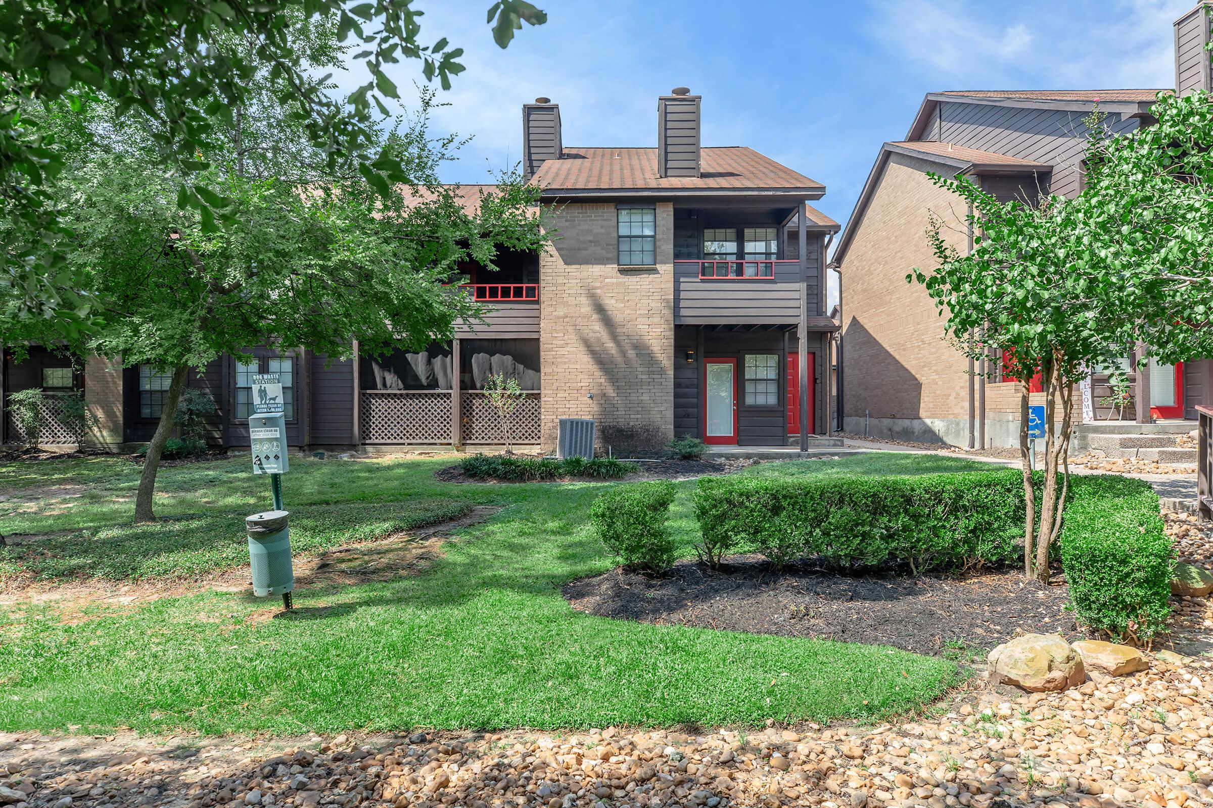 Two multi-story apartment buildings surrounded by greenery. In the foreground, well-maintained grass and shrubs are visible, along with decorative rocks. The apartment units feature balconies with red accents and a pathway leading towards the building. A trash bin is visible near the pathway.