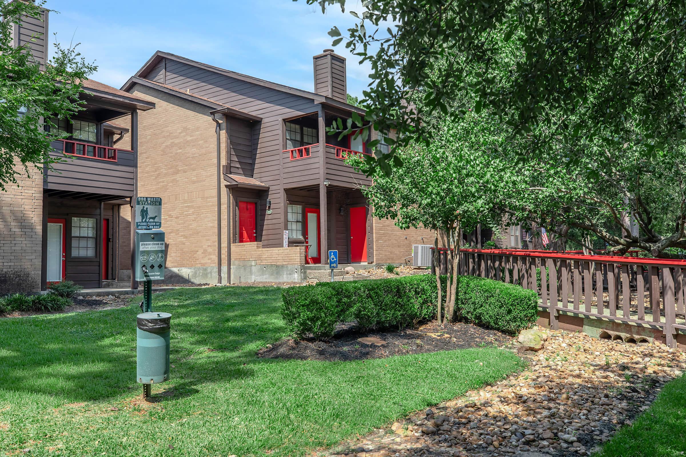Two-story apartment buildings with brown siding and red doors, set in a well-maintained landscape. Green lawn area with shrubs and a walking path made of rocks. A water meter can be seen in the foreground, along with a wooden railing that leads towards the entrance of one of the buildings. Clear blue skies above.
