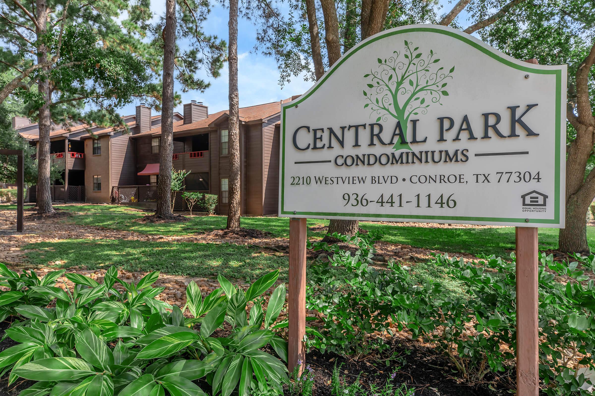 Sign for Central Park Condominiums, featuring the name prominently at the top. Below, it lists the address: 2210 Westview Blvd, Conroe, TX 77304, along with a contact number: 936-441-1146. The background shows a landscaped area with trees and shrubs, and condominium buildings in a residential setting.