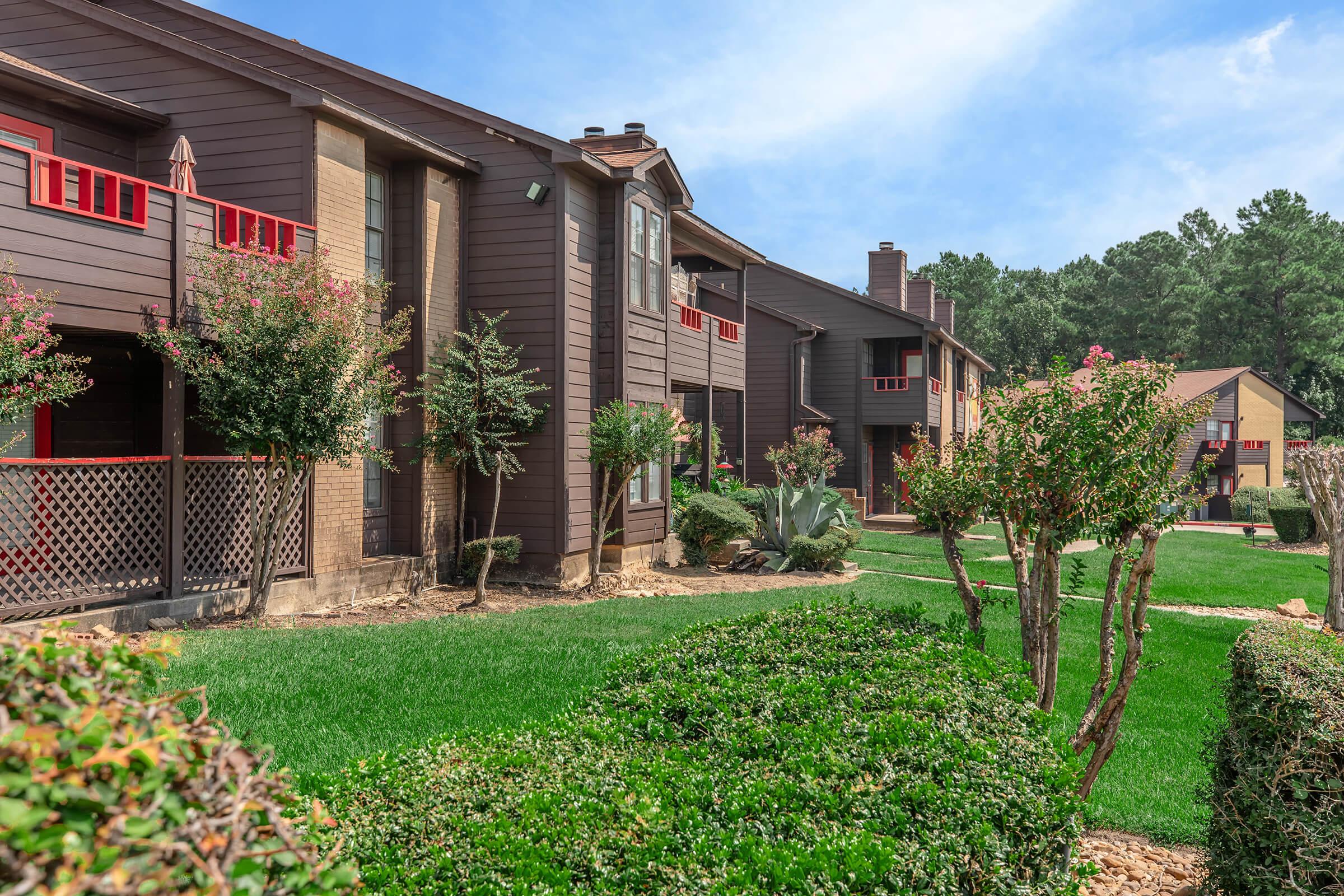 A view of a residential complex featuring dark wooden buildings with red accents. The landscape includes well-maintained green lawns, shrubs, and trees. The sky is clear and blue, indicating a sunny day. The architecture presents a modern yet cozy aesthetic in a tranquil setting.