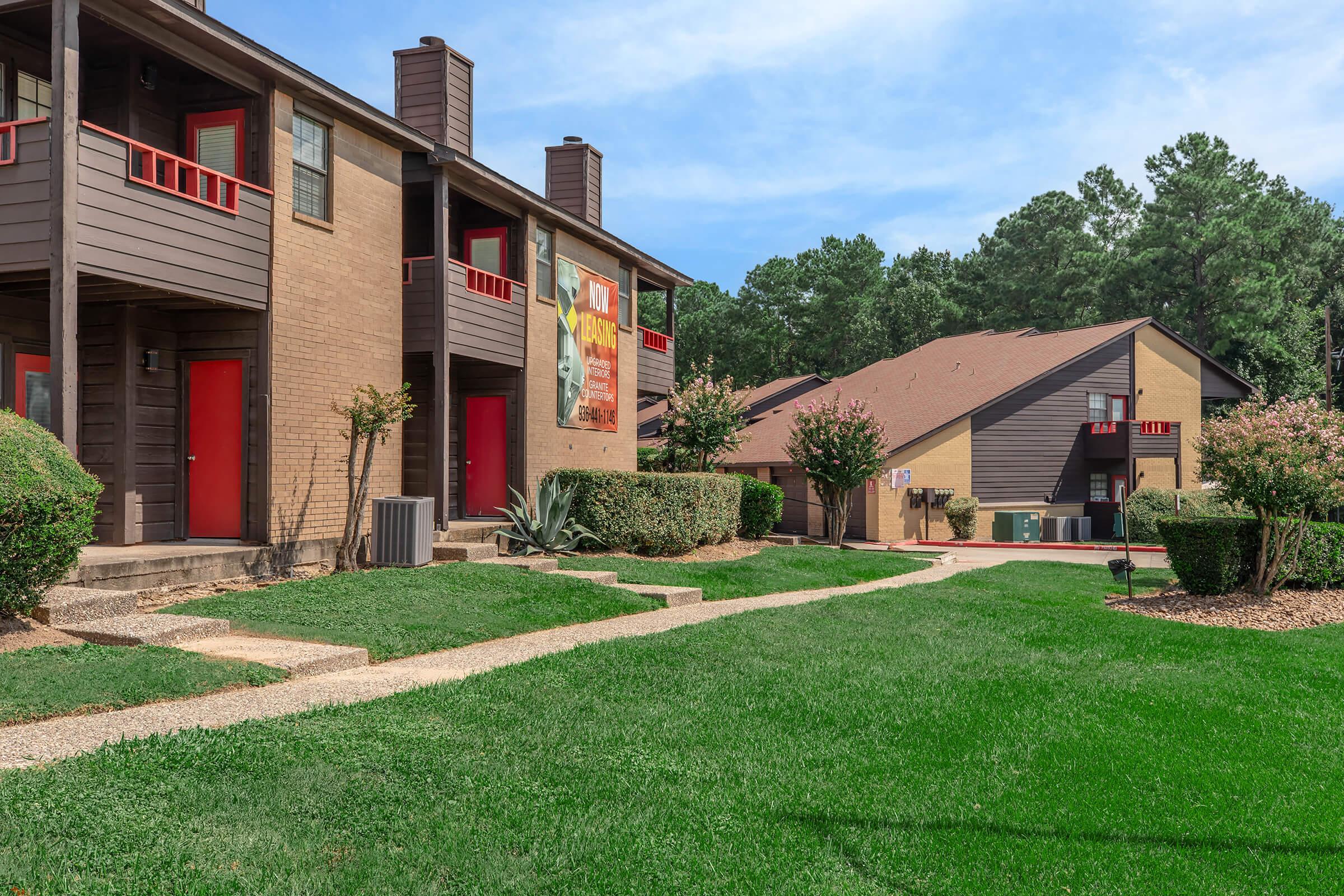 A residential complex featuring two, two-story apartment buildings. The foreground includes well-maintained lawns with pathways leading to the entrances. Each building has red doors and balconies, surrounded by ornamental bushes and trees. The sky is clear, providing a bright and inviting atmosphere.