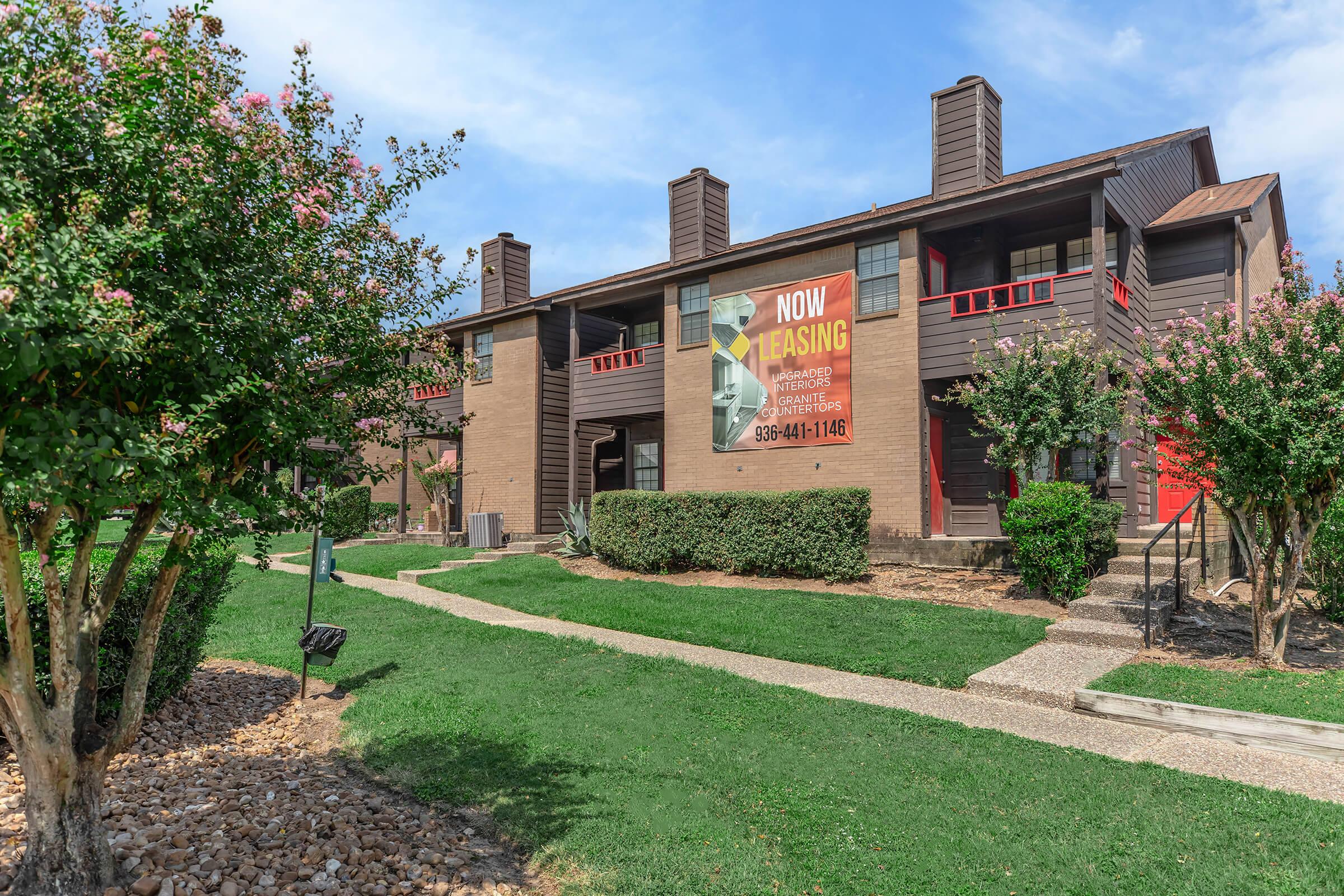 A two-story apartment building featuring a large "Now Leasing" banner on the front. The building is surrounded by neatly trimmed grass, flowering bushes, and a stone pathway leading to the entrances. There are several trees and landscaping elements enhancing the exterior appeal.