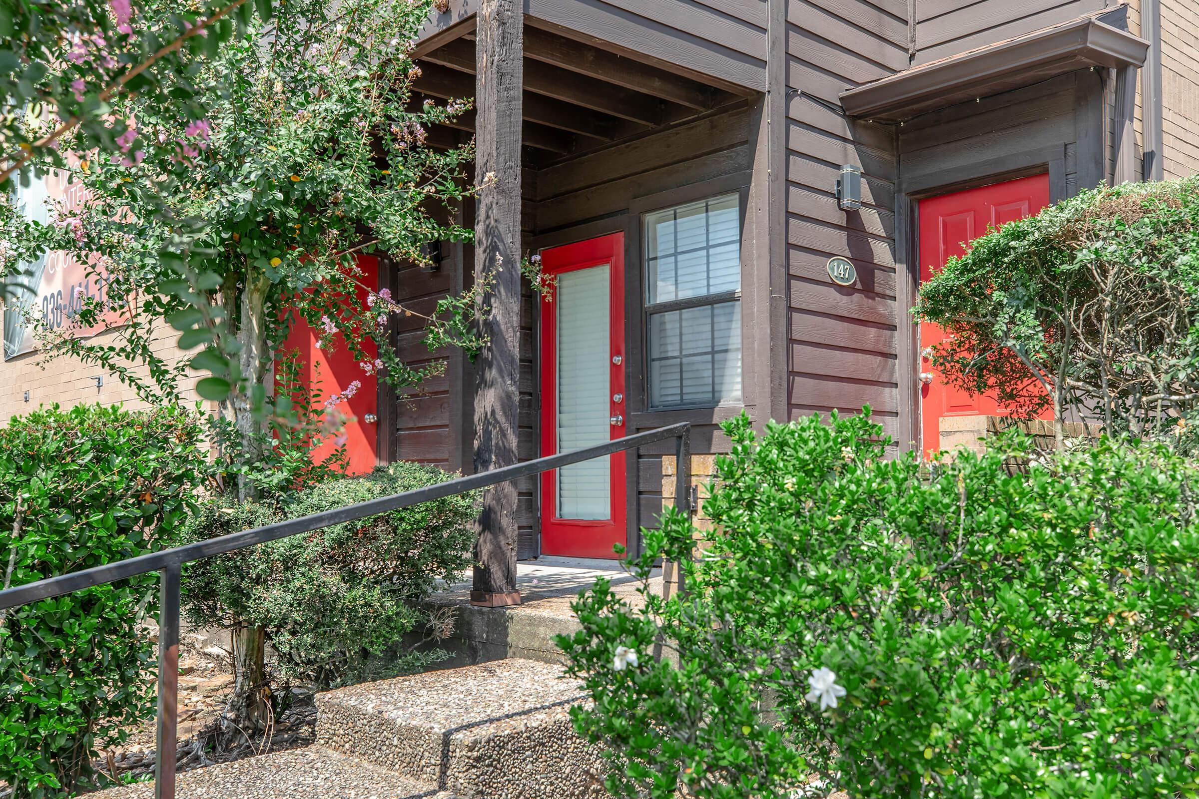 A brown wooden exterior of a two-story building with bright red doors, surrounded by well-trimmed green bushes. A staircase leads up to the entrance, with decorative plants on either side. The setting appears inviting and well-maintained.