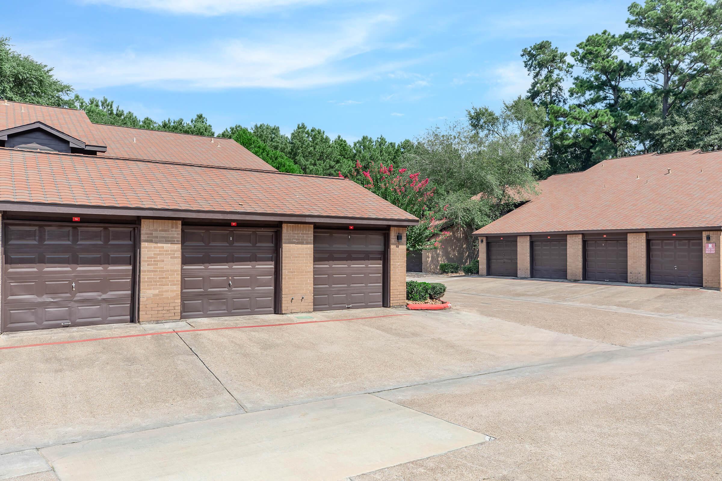 Row of brick garages with brown doors under a blue sky. The driveways are paved, and green trees and shrubbery are visible in the background. The scene is well-lit and appears to be part of a residential area or complex.