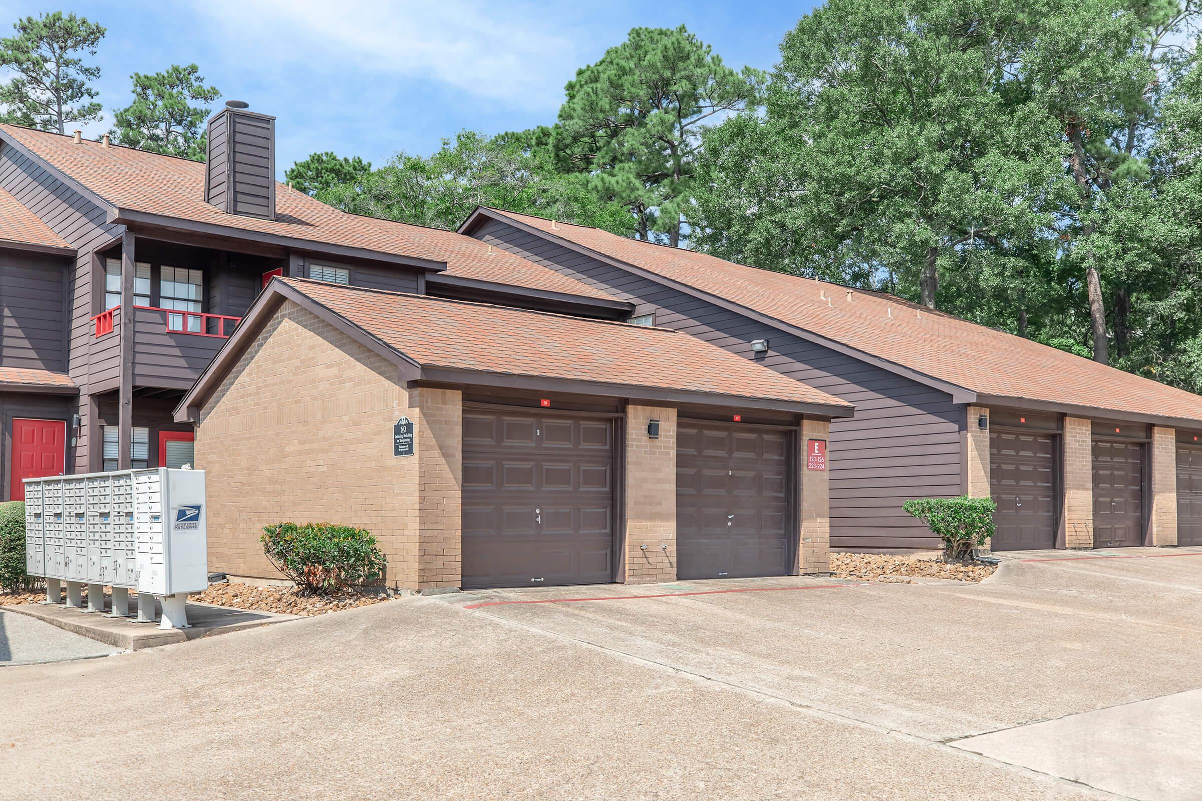 Exterior view of a residential building featuring multiple garage doors and a mailbox station. The structure has a brown wood siding and a sloped roof, surrounded by landscaped greenery.