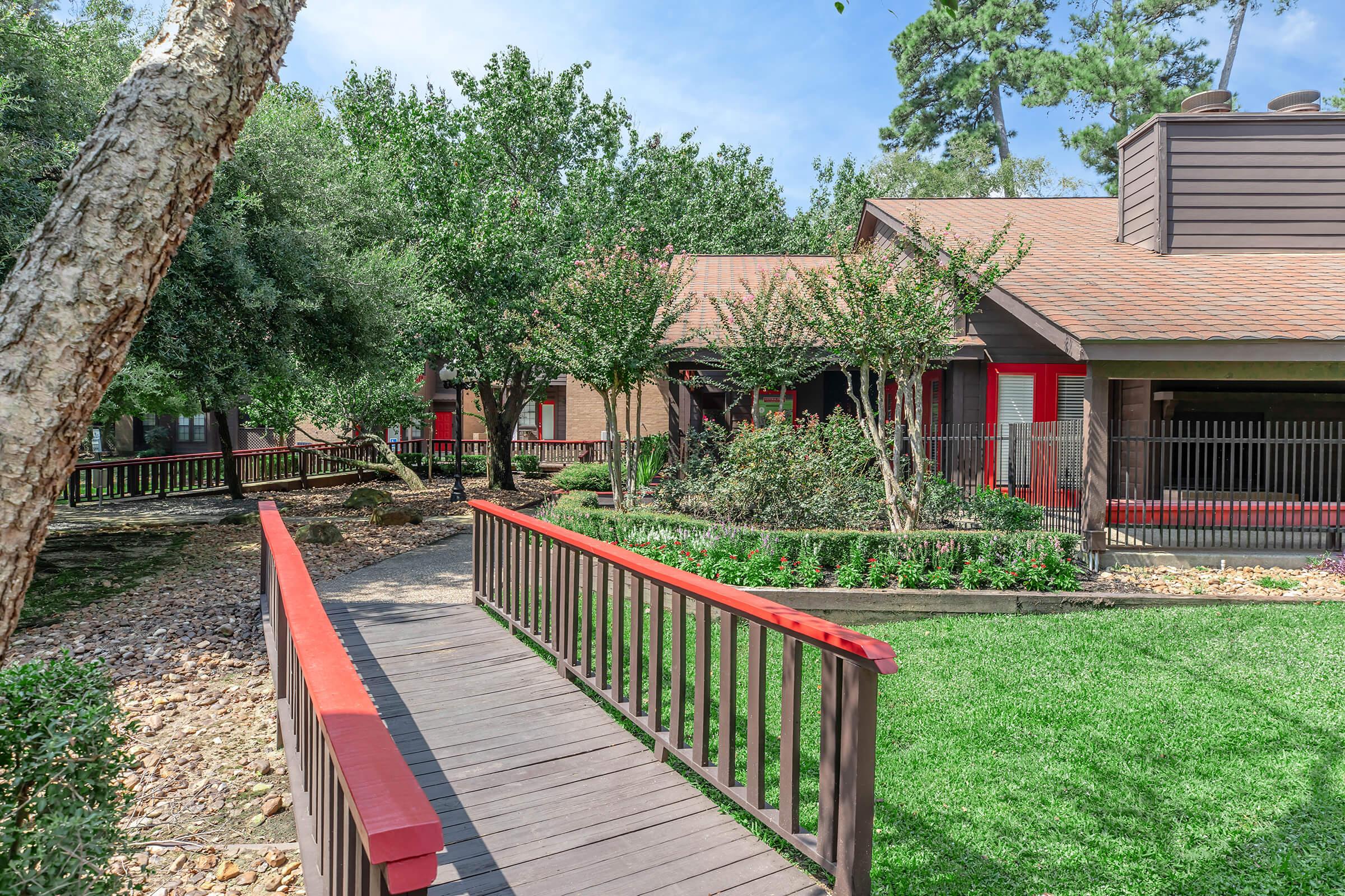 A peaceful outdoor scene featuring a walkway with a wooden railing leading through a green lawn surrounded by trees and shrubs. In the background, a wooden structure with red accents can be seen, enhancing the tranquil atmosphere of the landscape. Bright blue skies add to the sunny ambiance.