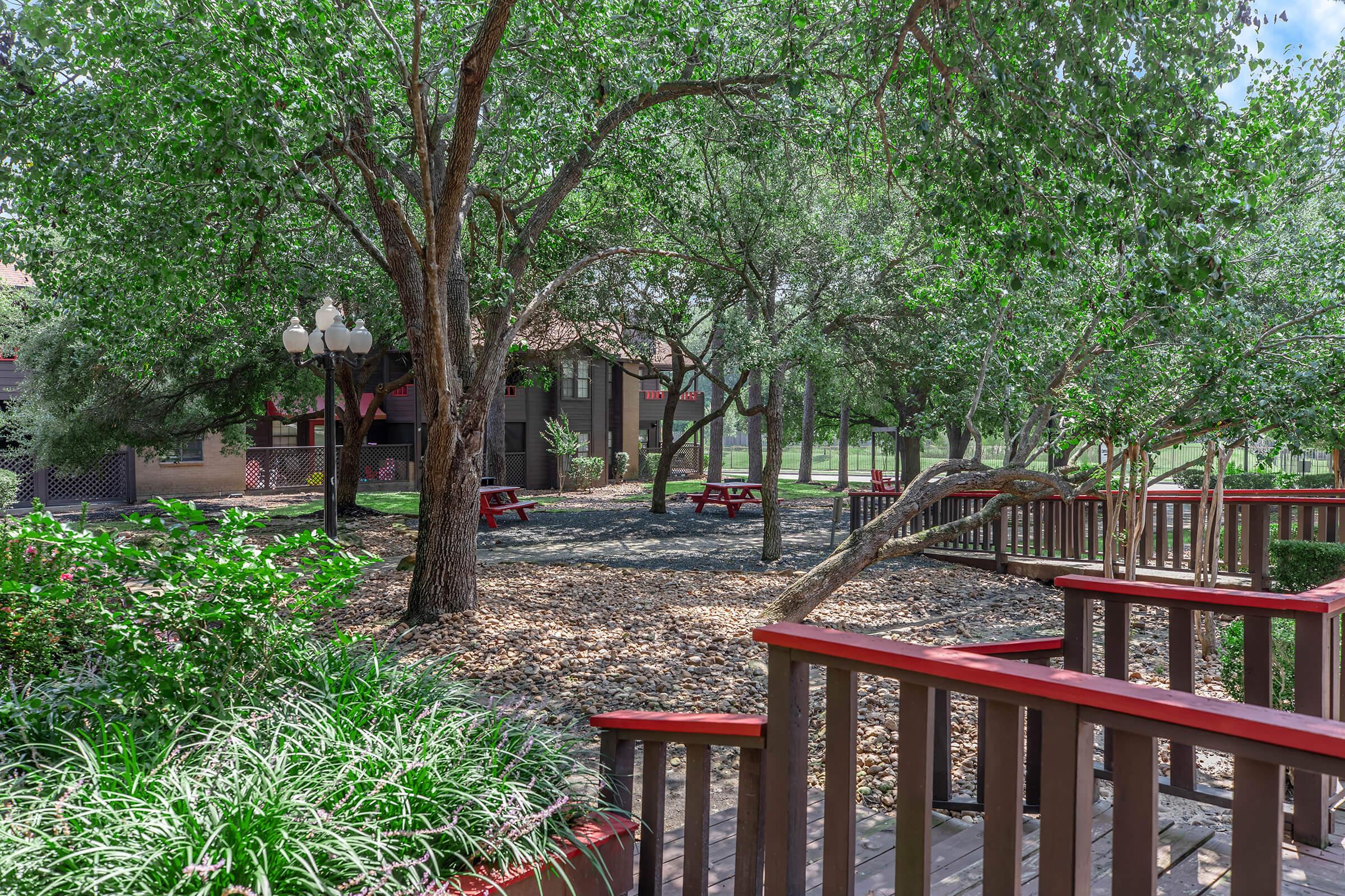 A serene outdoor area featuring lush green trees and shrubs, a wooden walkway with red railings, and picnic tables in the background. Bright sunlight filters through the leaves, creating a peaceful atmosphere, ideal for relaxation or outdoor gatherings.