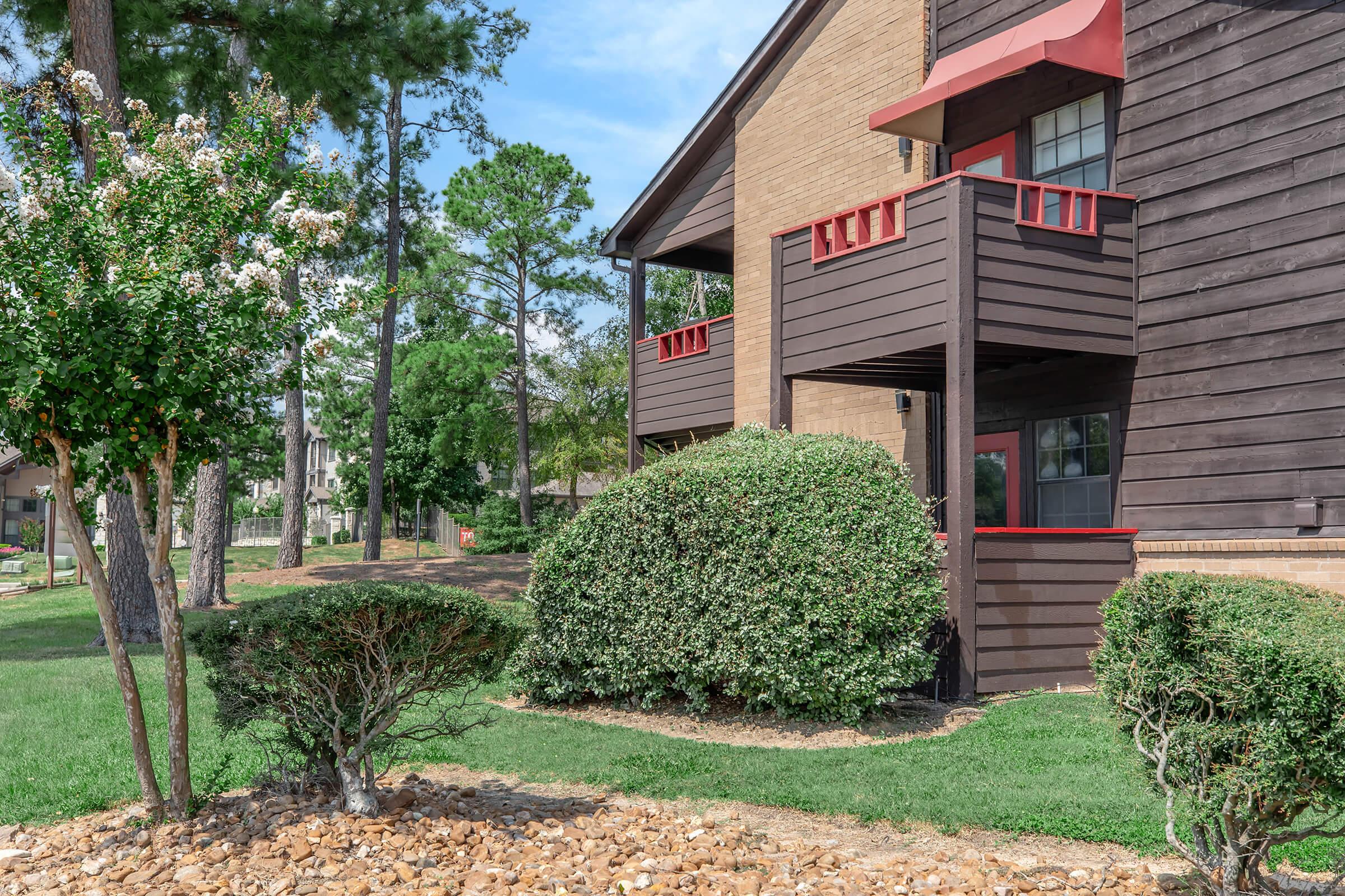 A view of a well-maintained residential area featuring a brown wooden building with red accents. There are neatly trimmed bushes and a small garden area with decorative stones. Trees provide shade in the background under a clear blue sky.